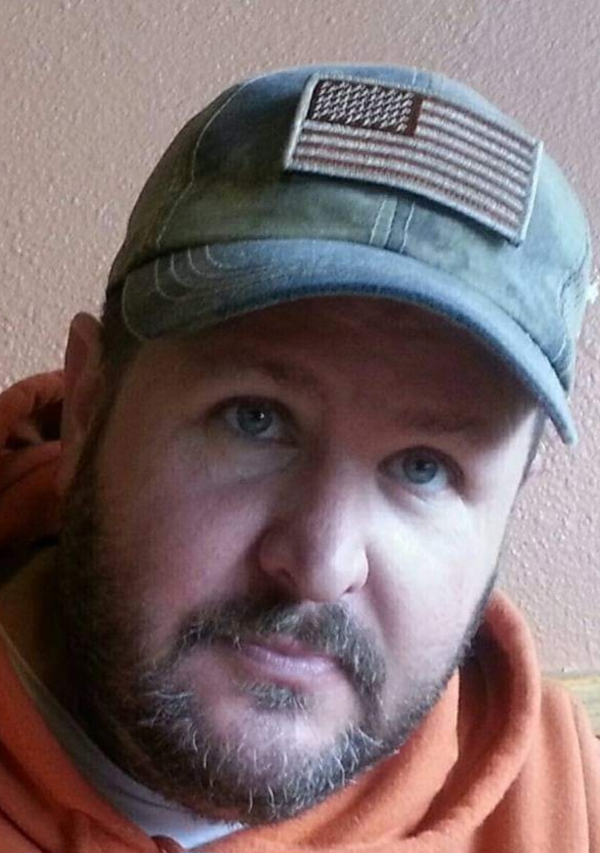Close-up of a man wearing a camouflage cap with an American flag patch, and an orange hoodie, against a plain wall.