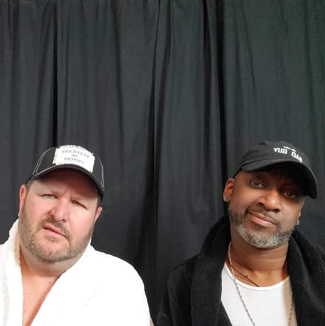 Two men pose in front of a black curtain, one wearing a white shirt and cap, the other in a black cap and jacket.