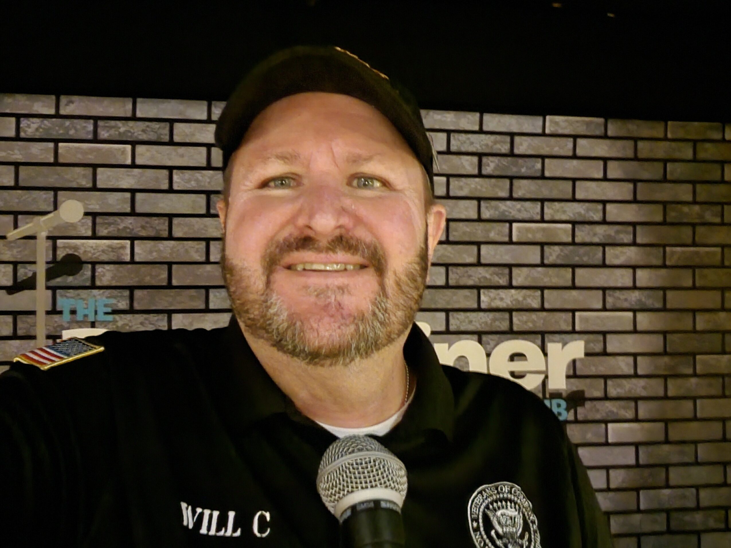 Man with a beard wearing a police uniform and cap, smiling, standing in front of a brick wall background.