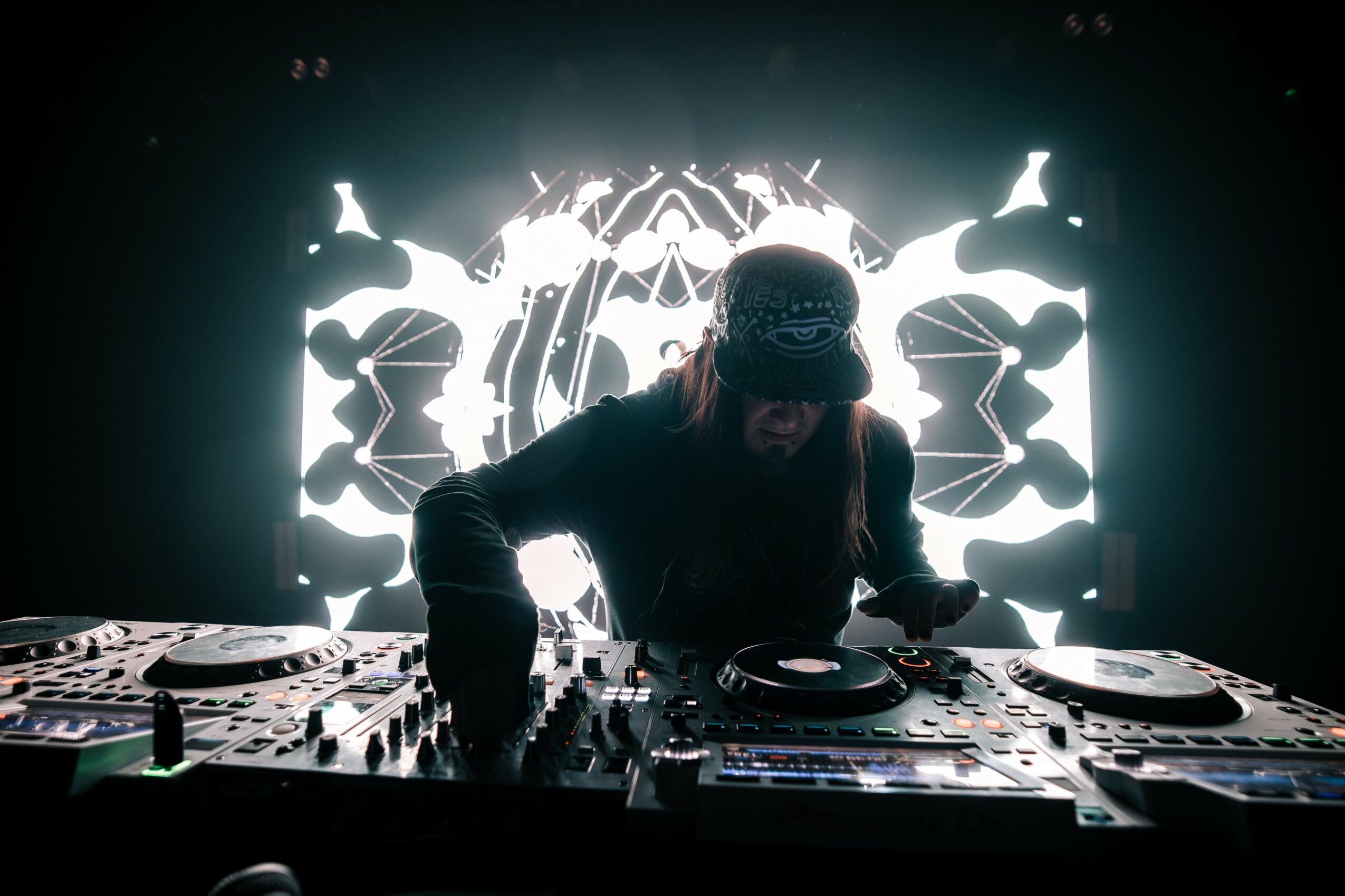 DJ wearing a cap, performing with equipment in front of a bright, symmetrical light display behind him.