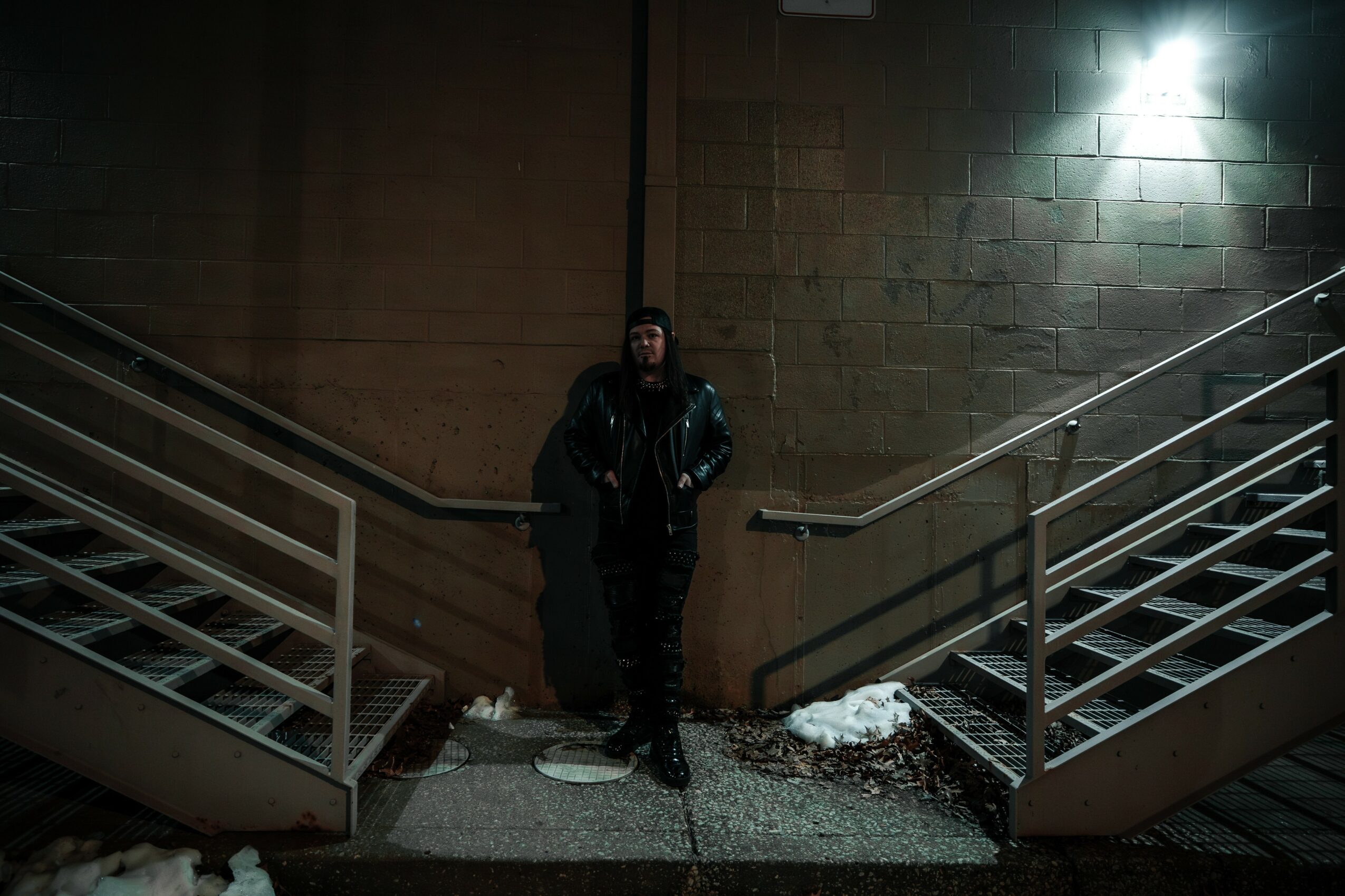 Person standing between two staircases outside at night, with snow on the ground and a brick wall background.