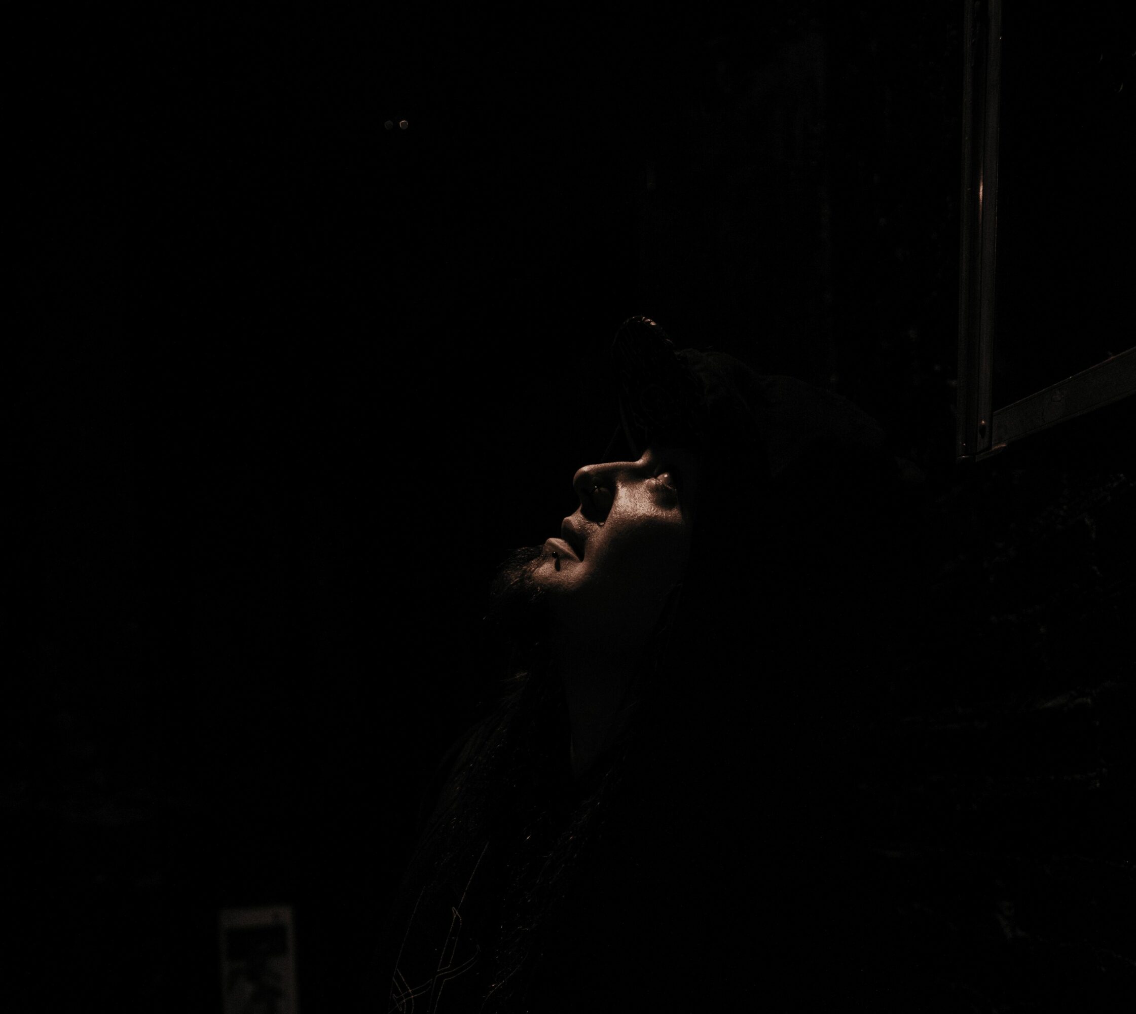 Person with face illuminated in dark environment, looking upward, hand on forehead, low light, reflective surface nearby.