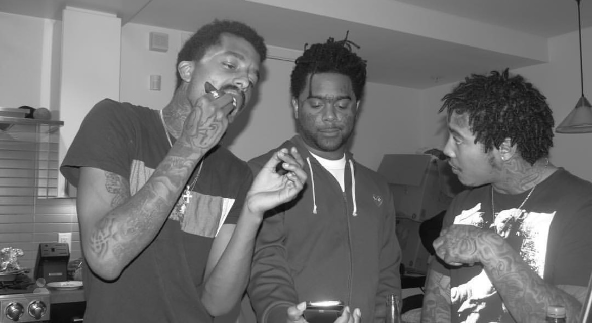 Three young men in a kitchen, one looking at a phone, one with hand near mouth, another looking at the phone.
