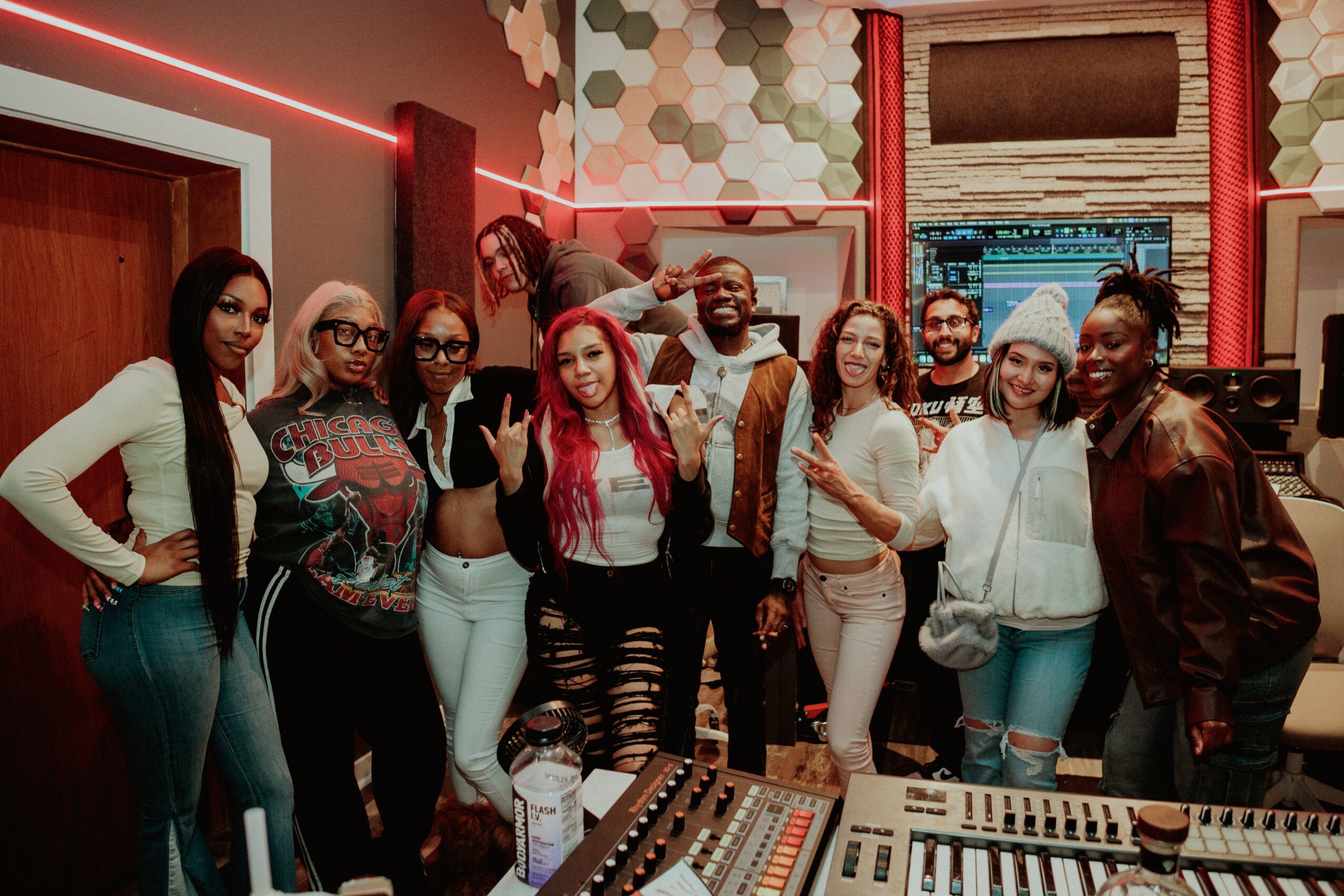 Group of ten people in a recording studio, some making peace signs, with audio equipment and colorful wall decor.