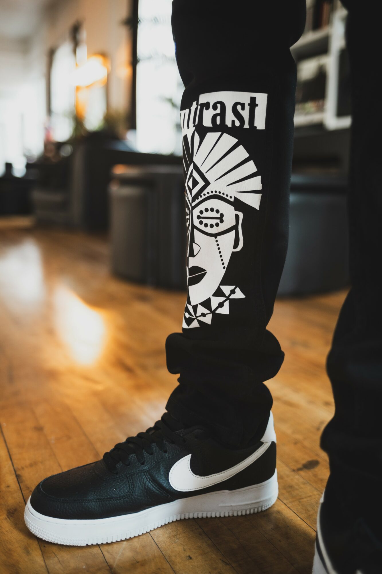 Person wearing black pants with graphic design and black Nike sneakers standing on wooden floor indoors.