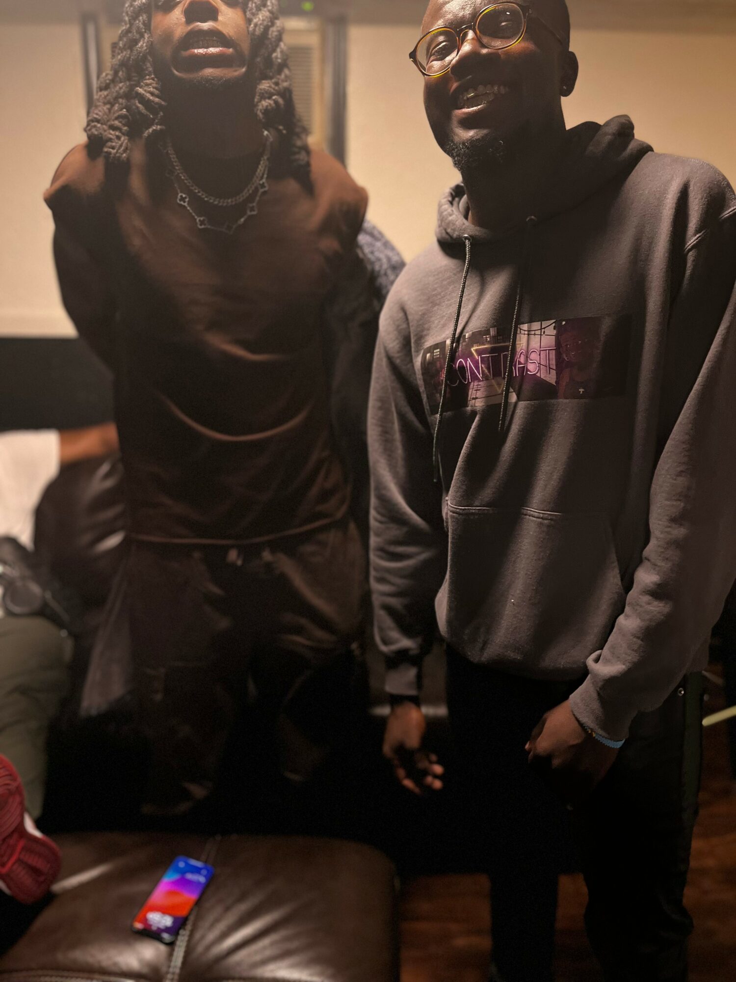 Two men standing indoors, one with long hair and glasses, the other with short hair and a hoodie, smiling.