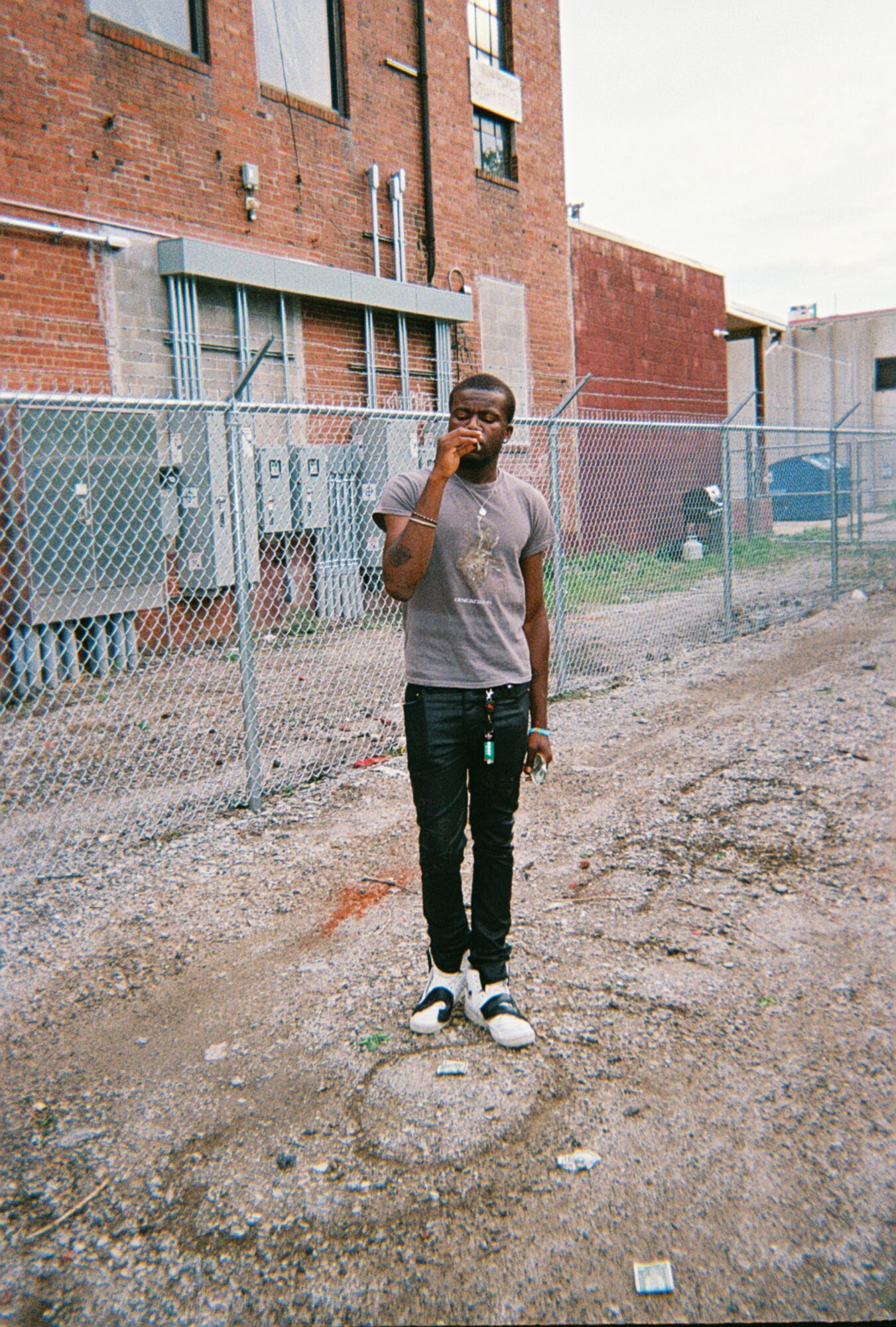 Young man standing on gravel ground near a chain-link fence with buildings in the background.