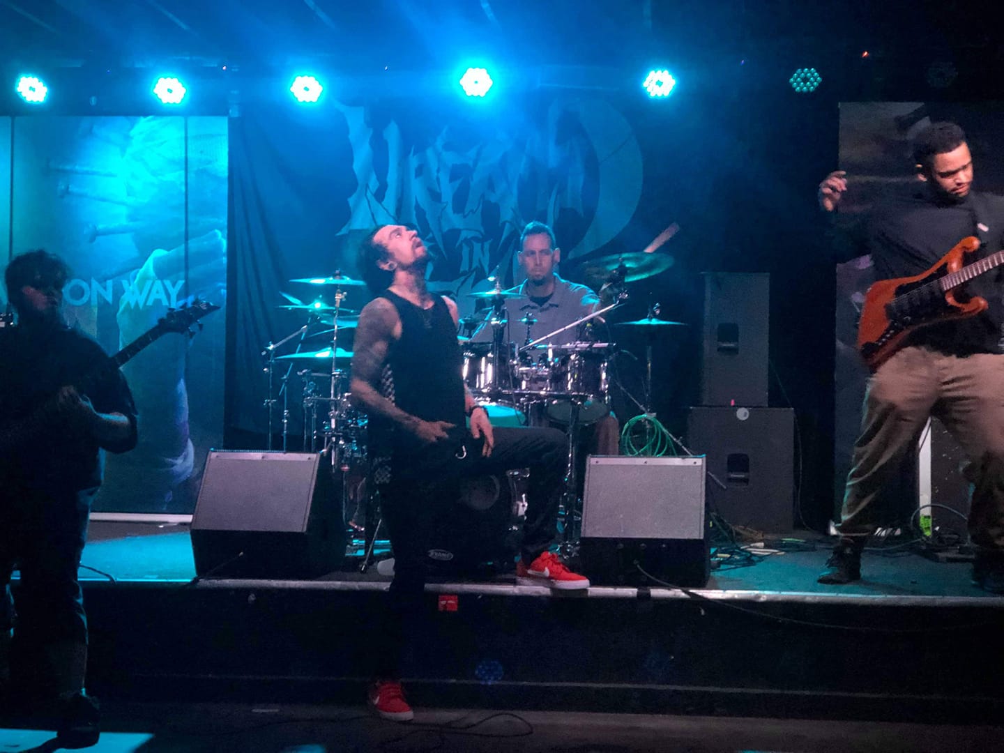 Band performing on stage with musicians playing guitar and drums, blue stage lighting, and a screen in the background.