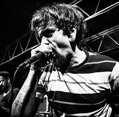 Man singing into microphone on stage, wearing striped shirt, with stage lights and structure in background.