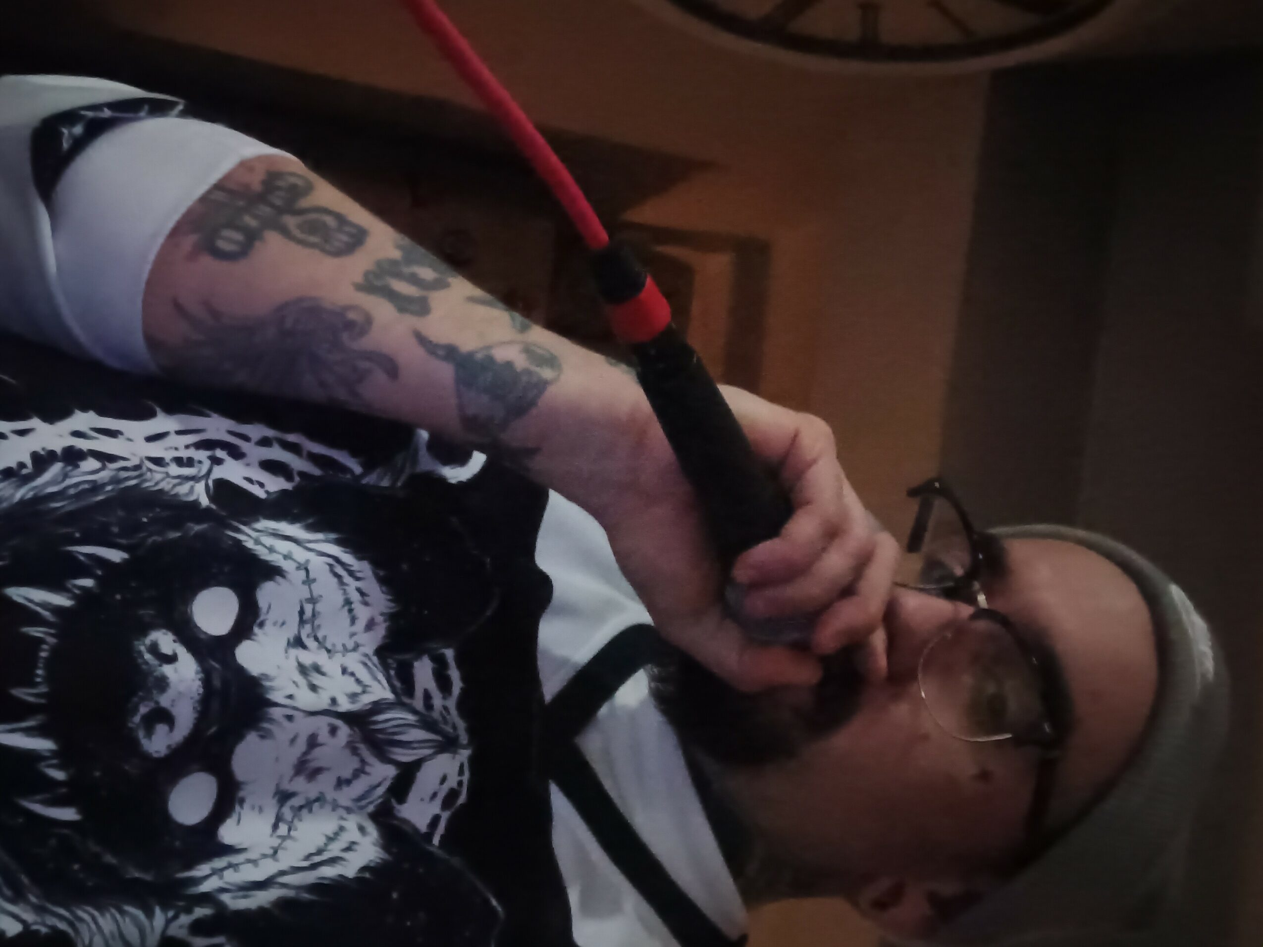 Person with tattoos on arm holding a red and black object near face, wearing glasses and a hat, indoors.