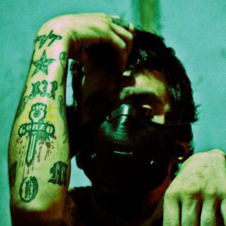 Person with tattoos on arm, wearing a black mask, holding hair, against a green background.