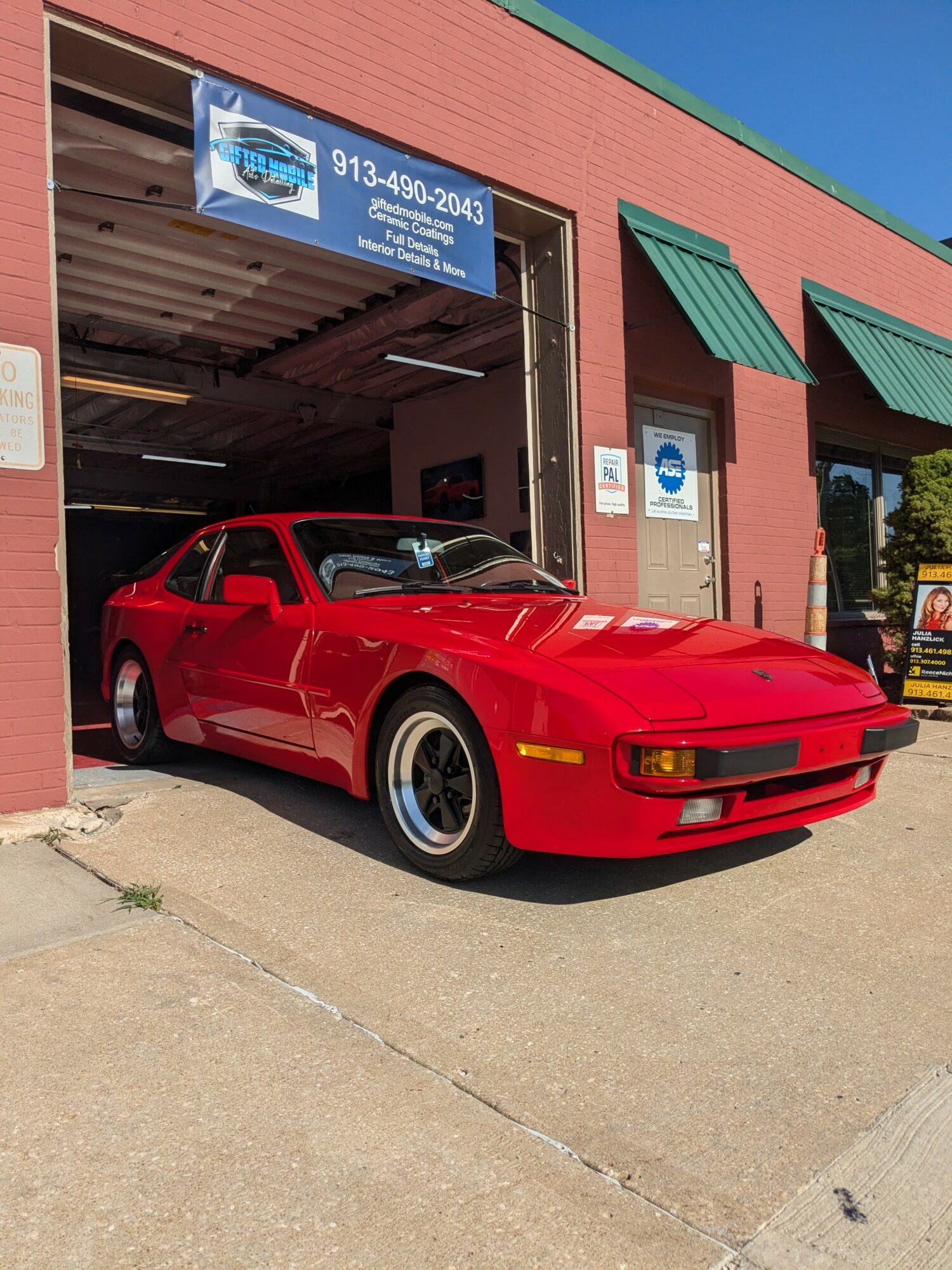 Red sports car parked outside a garage with a blue sign, green awnings, and a pink building, under a clear blue sky.