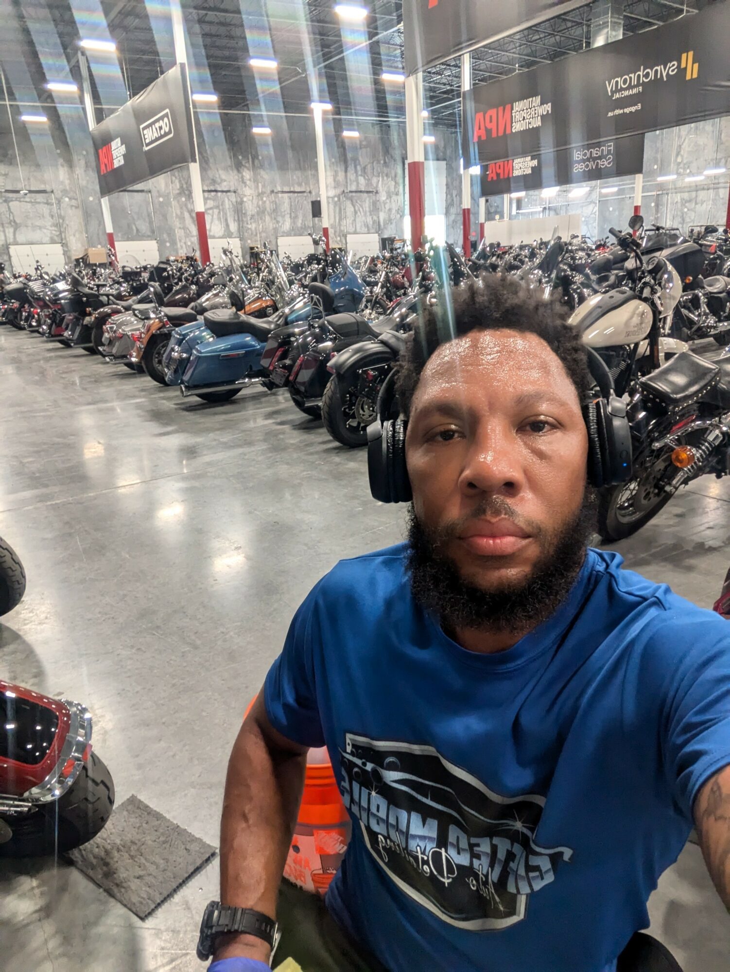 Man with headphones taking a selfie inside a motorcycle showroom with many motorcycles in the background.
