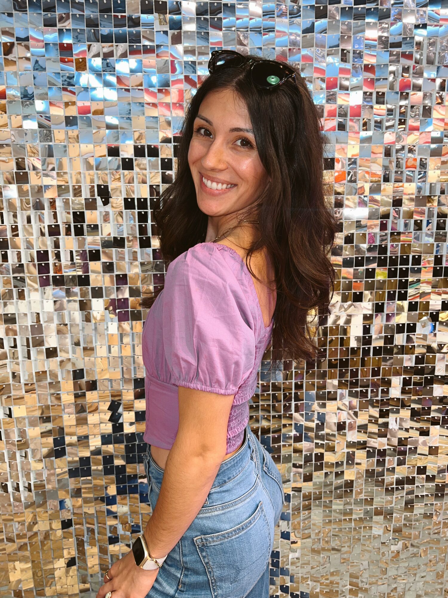 Smiling woman with long dark hair in a pink top and jeans, standing in front of a reflective mosaic wall.