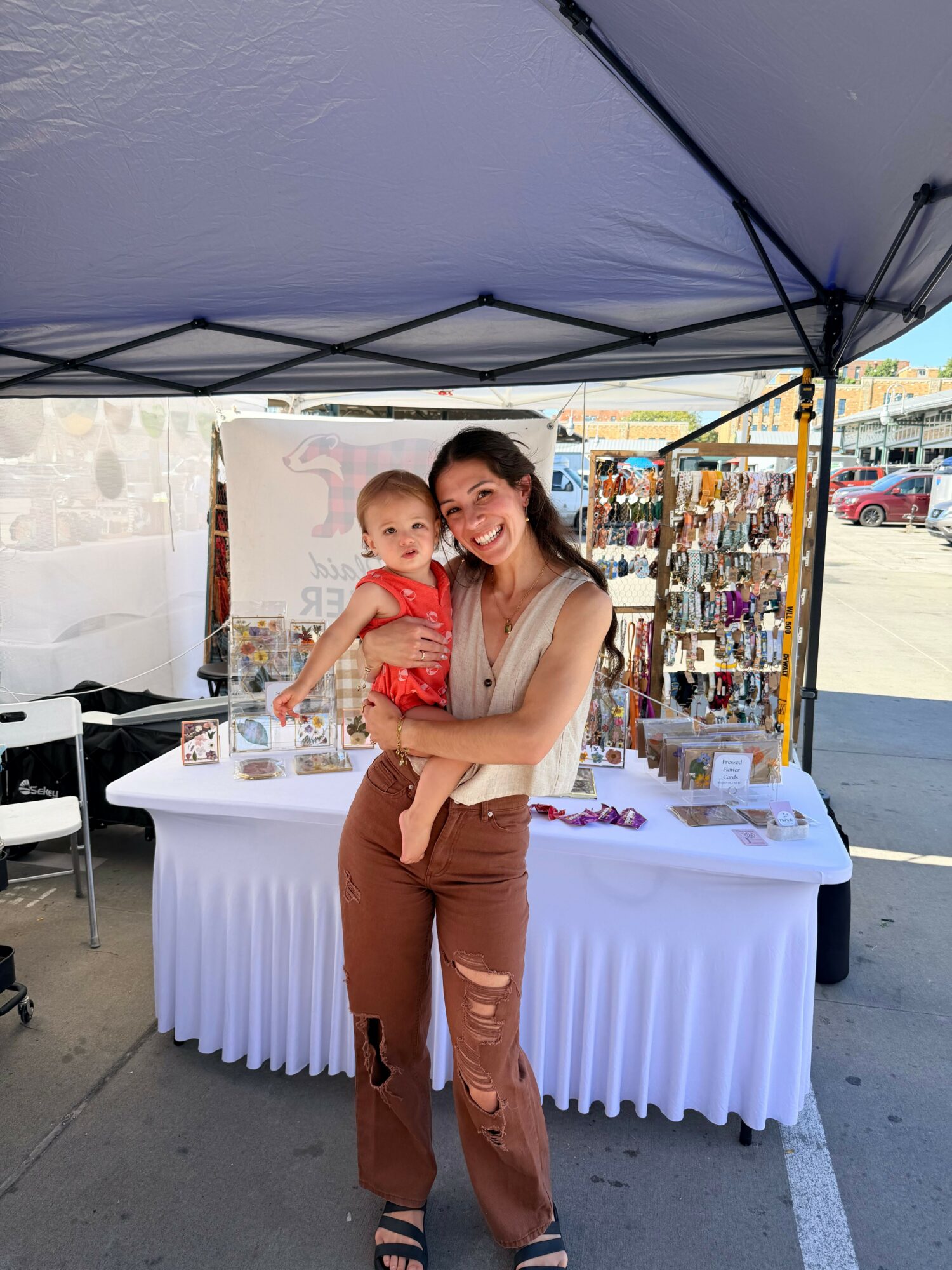 Woman holding a child under a large umbrella at an outdoor event, with tables and booths in the background.