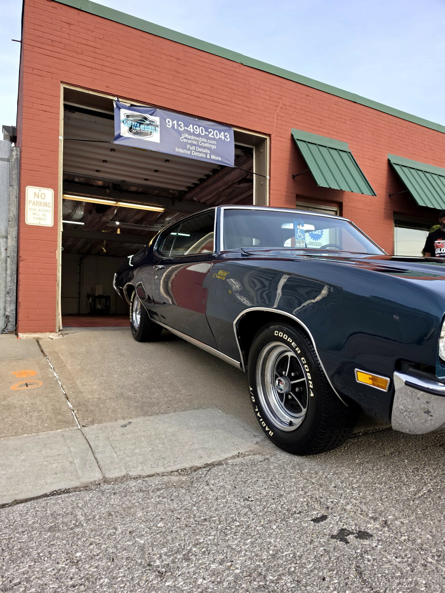 Black vintage car partially outside a brick building with green awnings, garage door open, and a sign above.