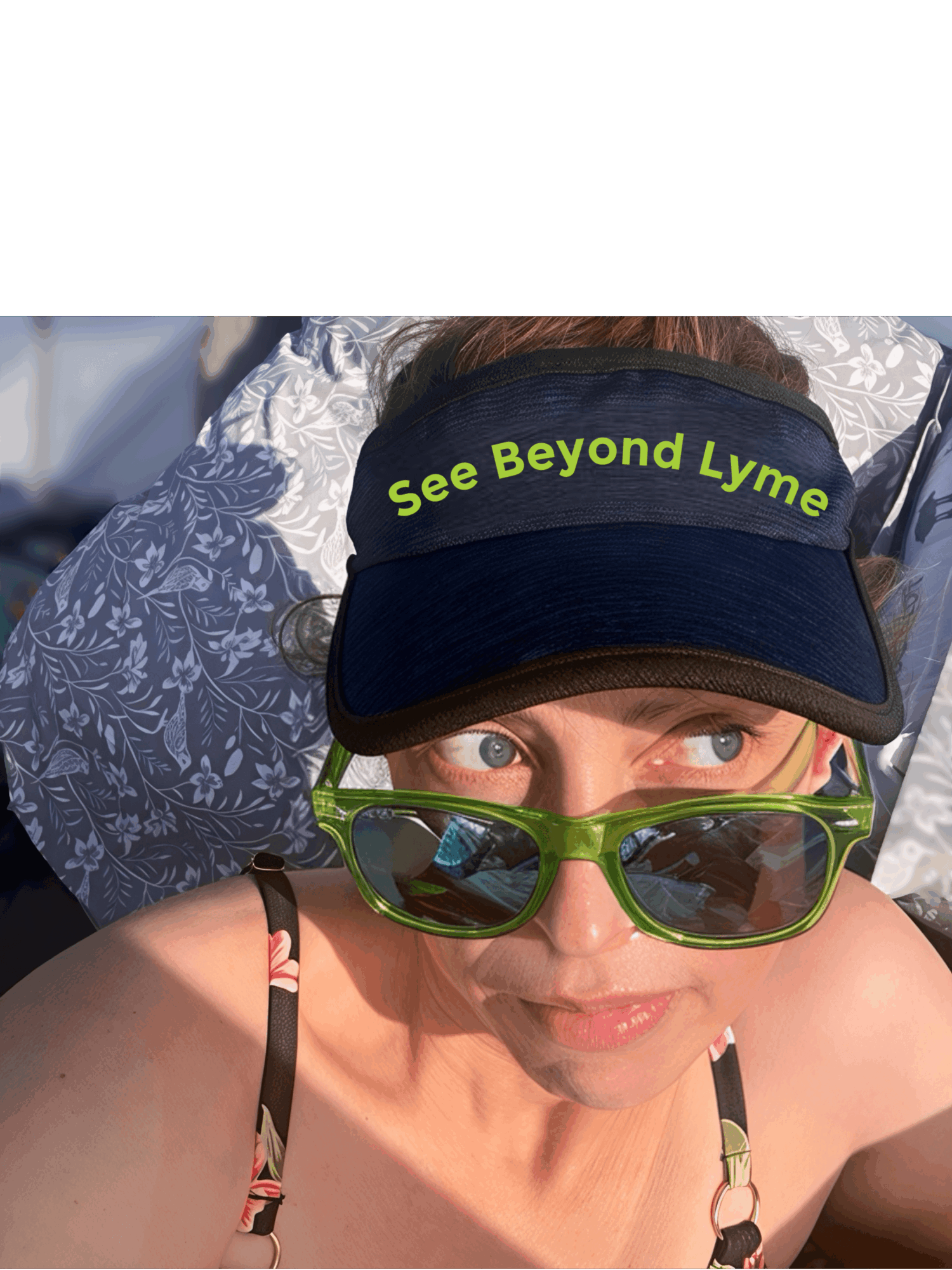 Woman wearing a visor with the text 'See Beyond Lyme' and green sunglasses, looking up at the camera.