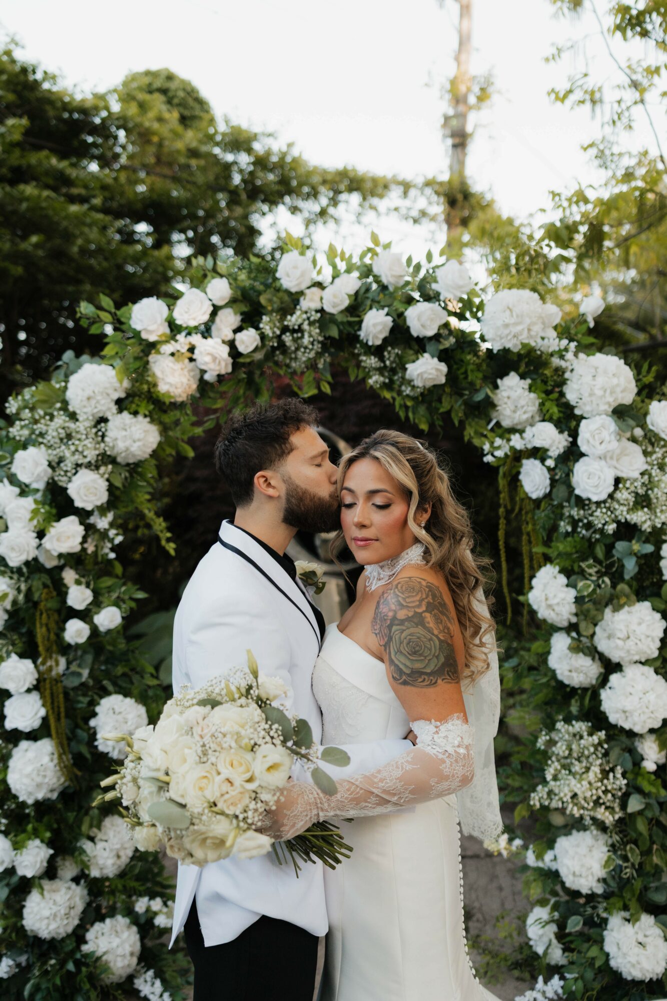 Bride and groom stand close under a floral arch, bride holding a bouquet, groom in a white suit, outdoor setting.