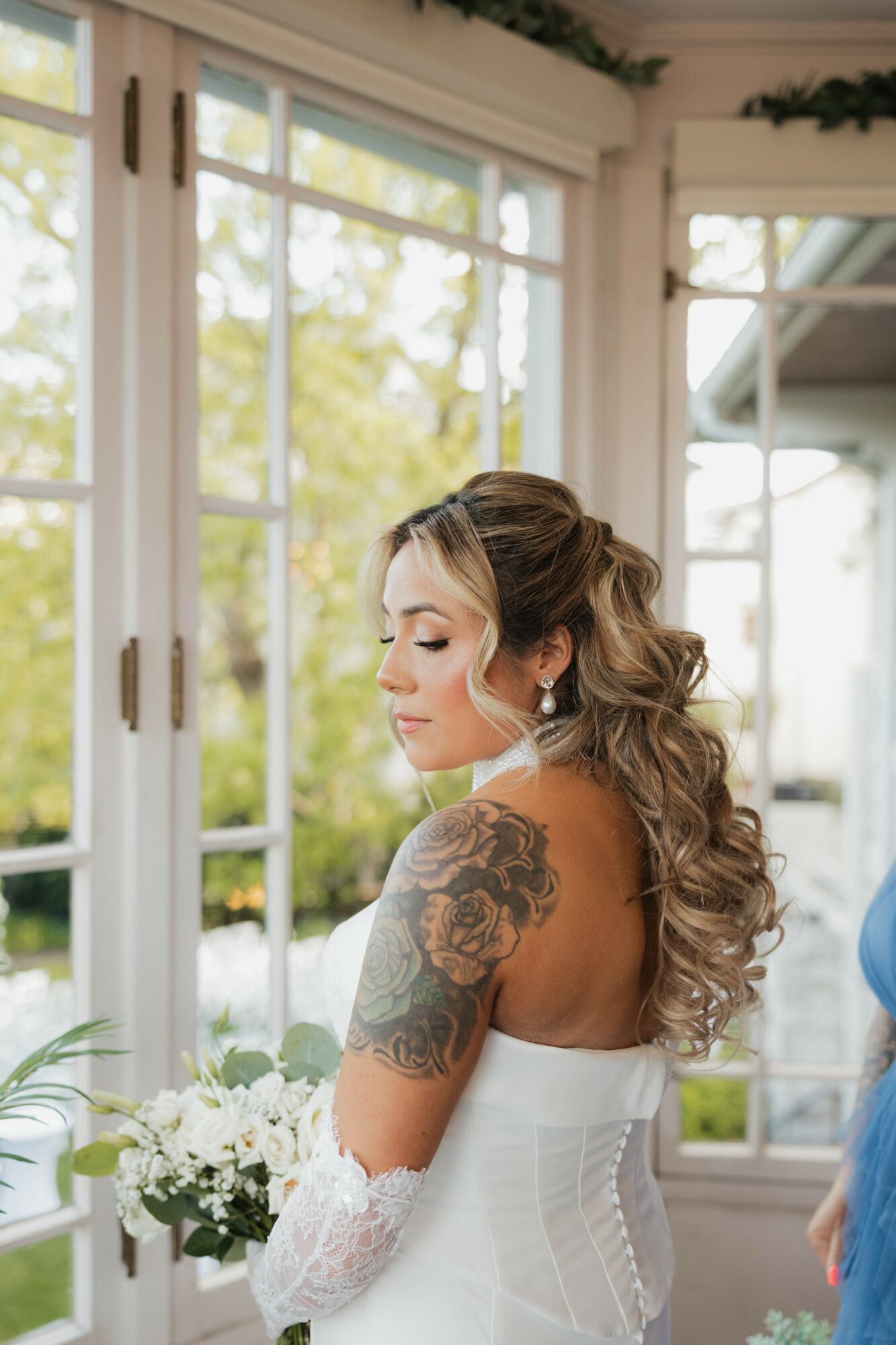 Woman with long curly hair and floral tattoo on shoulder, holding a bouquet, standing near large window with natural light.