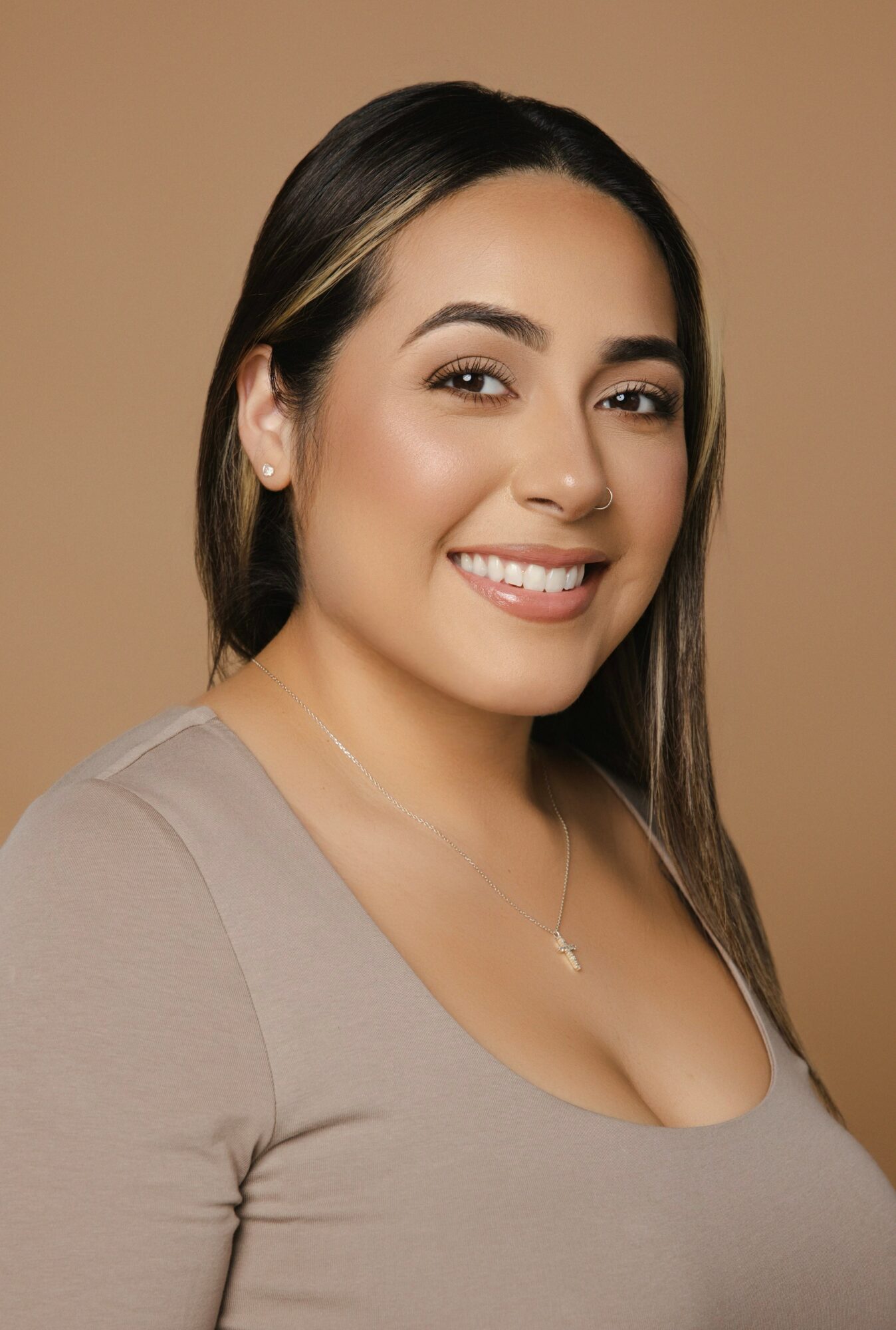 Smiling woman with long dark hair, wearing a beige top and necklace, against a plain background.
