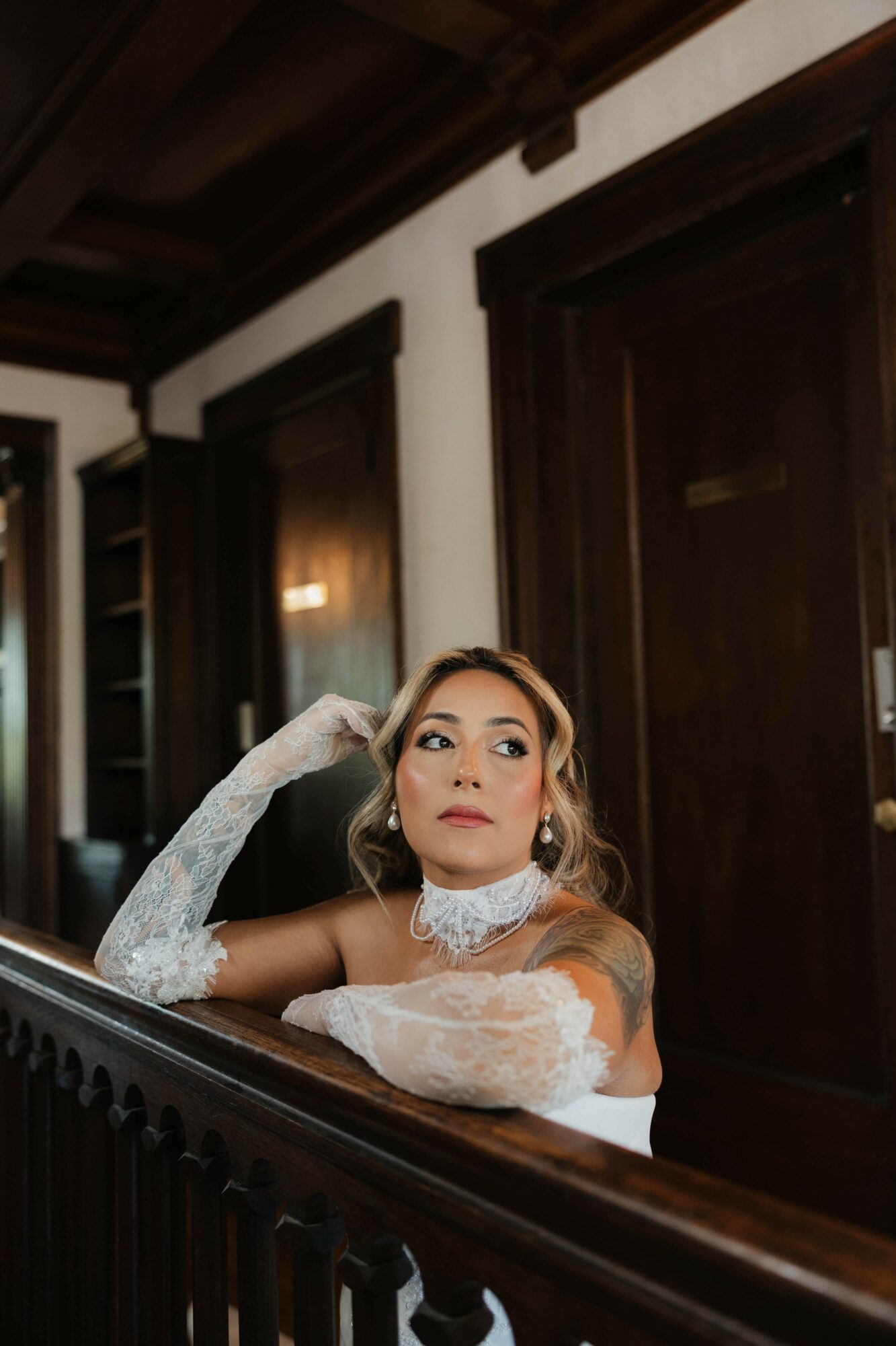 Woman with makeup and jewelry, resting her chin on her hand, in a room with dark wooden doors and paneling.