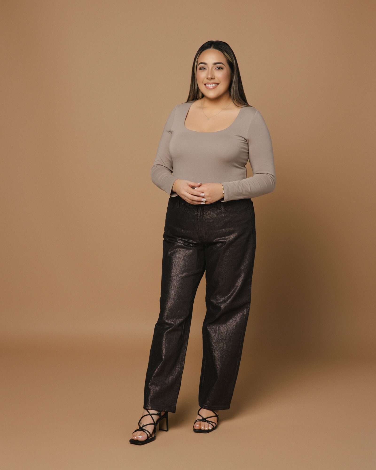Woman standing with hands clasped, smiling, wearing a gray long-sleeve top and black pants, against a beige background.