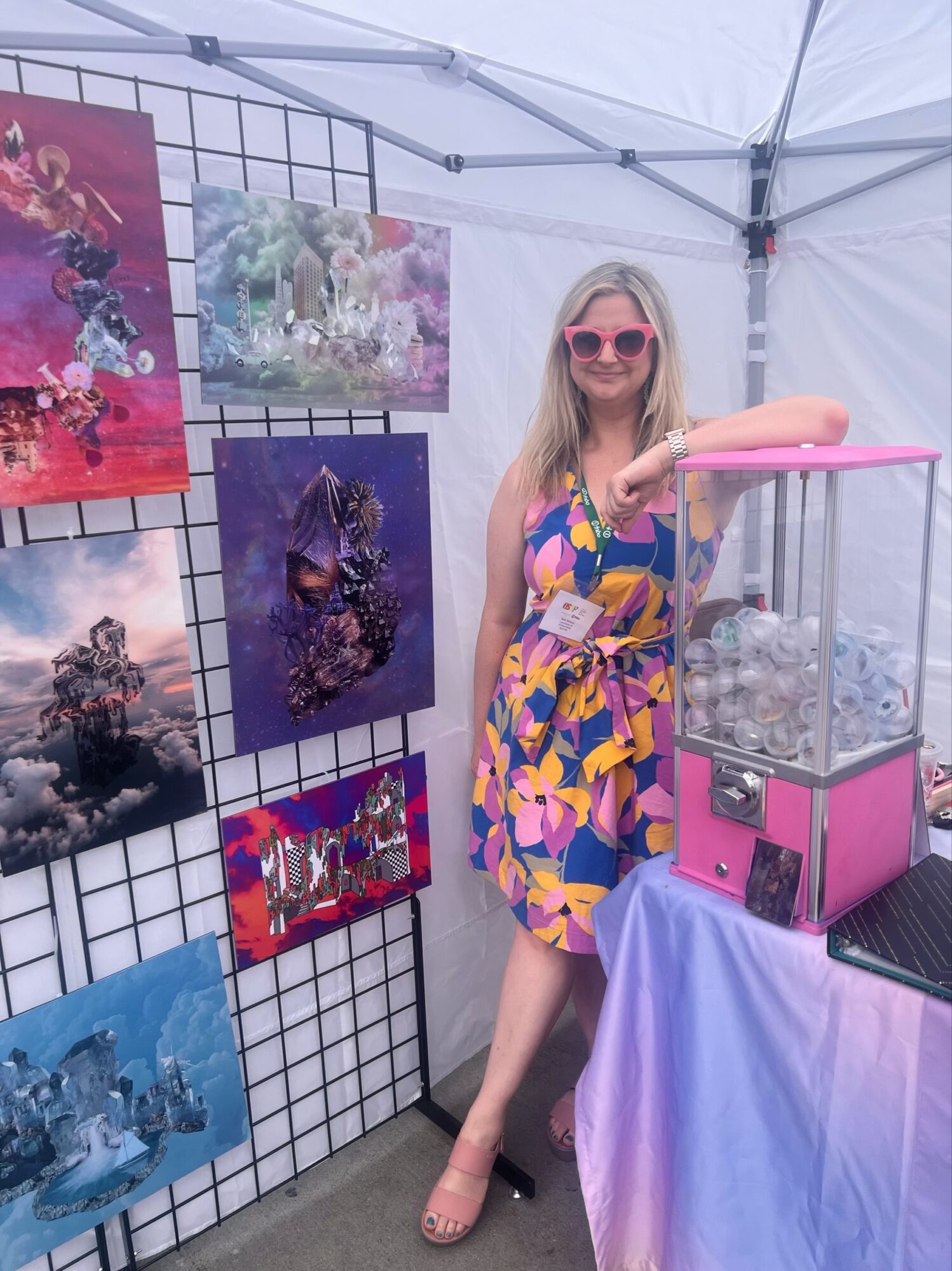 Woman in colorful dress and sunglasses standing next to a pink raffle drum inside a tent, with artwork displayed on a grid behind her.