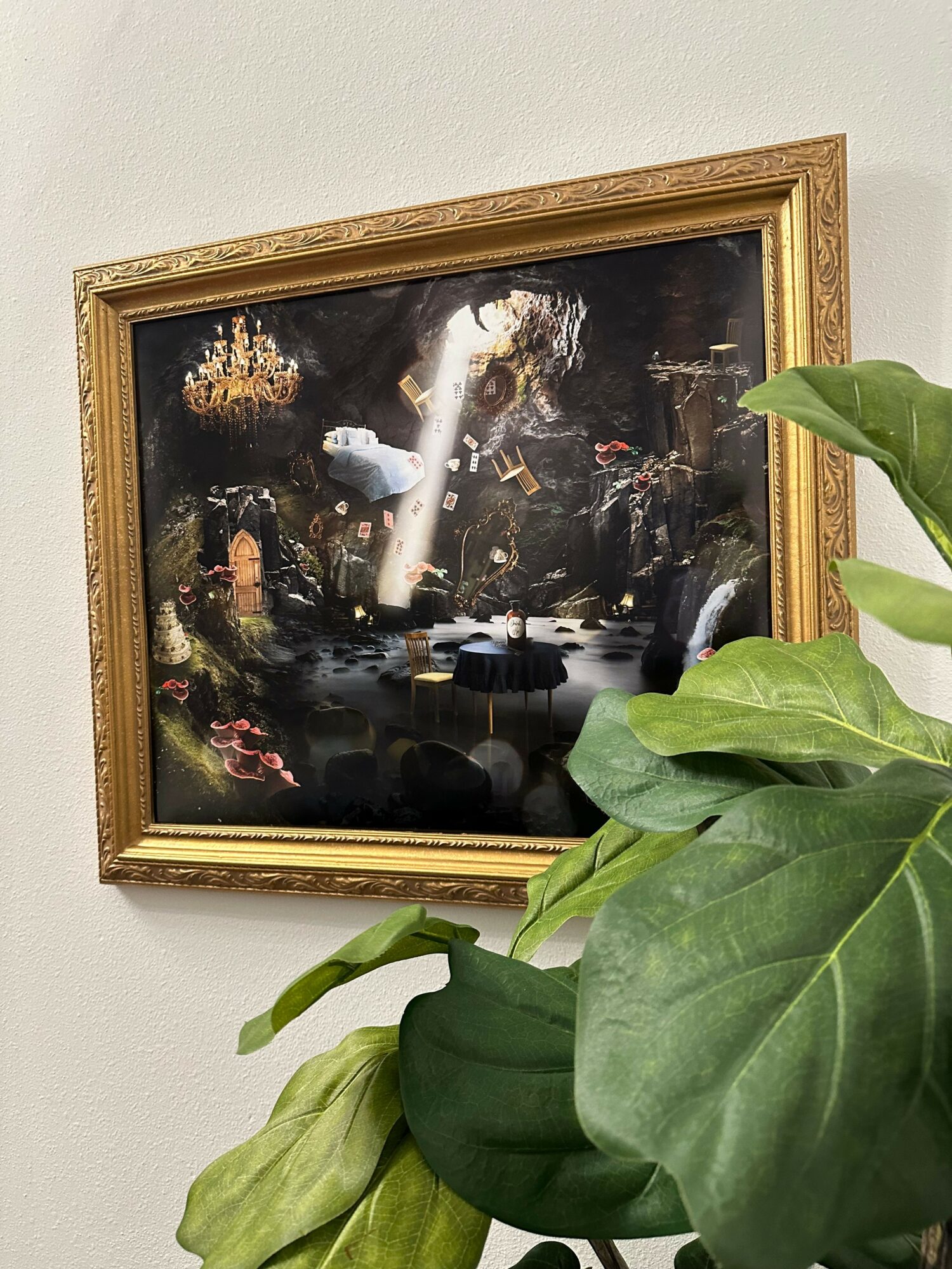Framed painting with dark colors, partially obscured by large green leaves in the foreground.