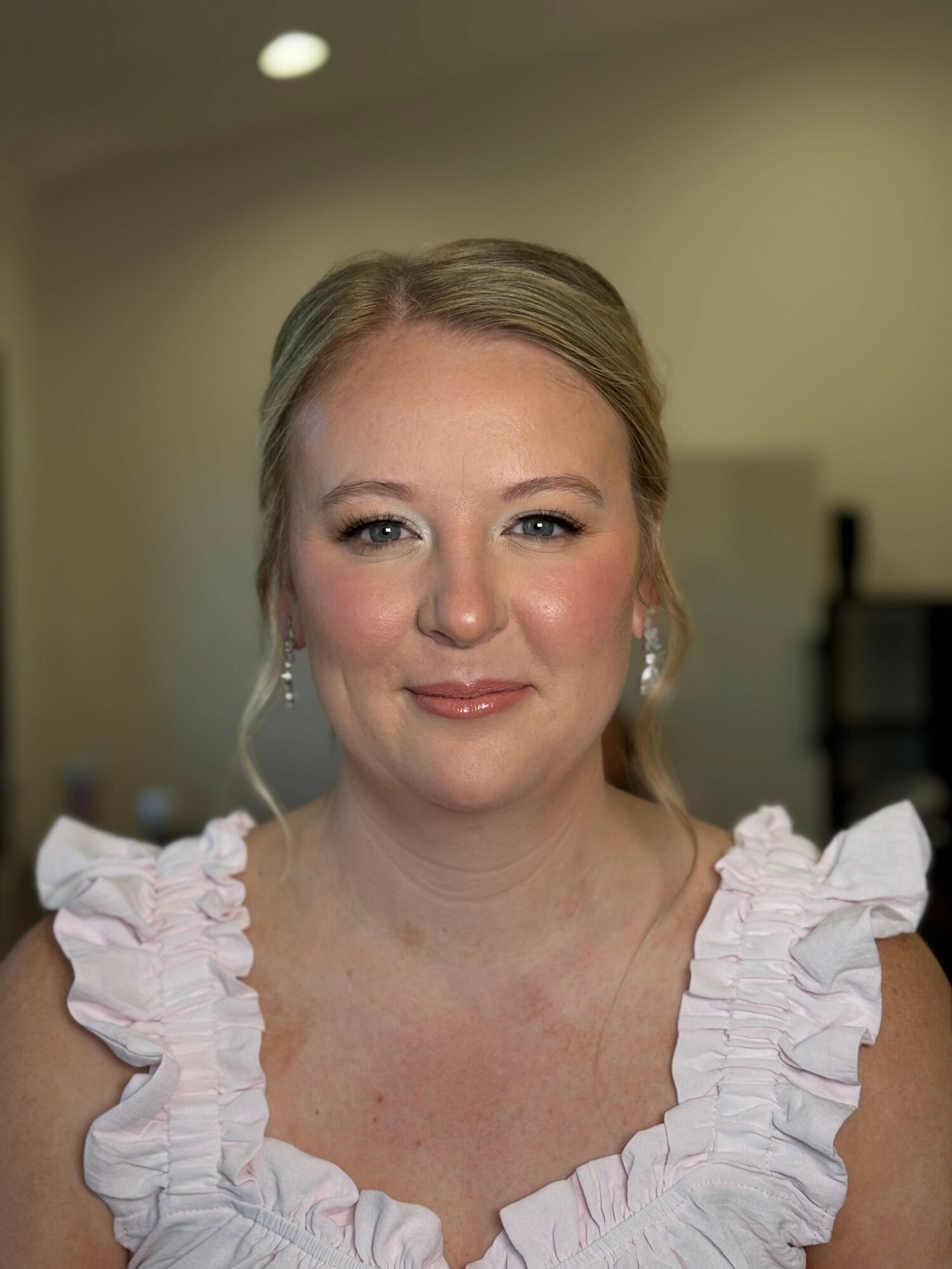 Portrait of a woman with blonde hair, wearing a ruffled white dress, smiling softly, indoors with blurred background.