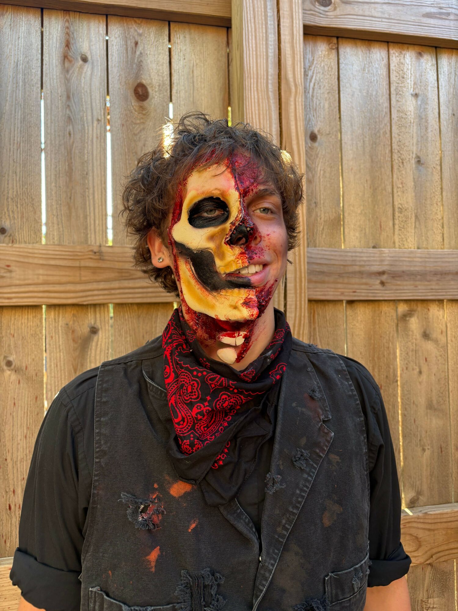 Person with face painted in bright colors, wearing a black jacket and red bandana, standing against a wooden fence.