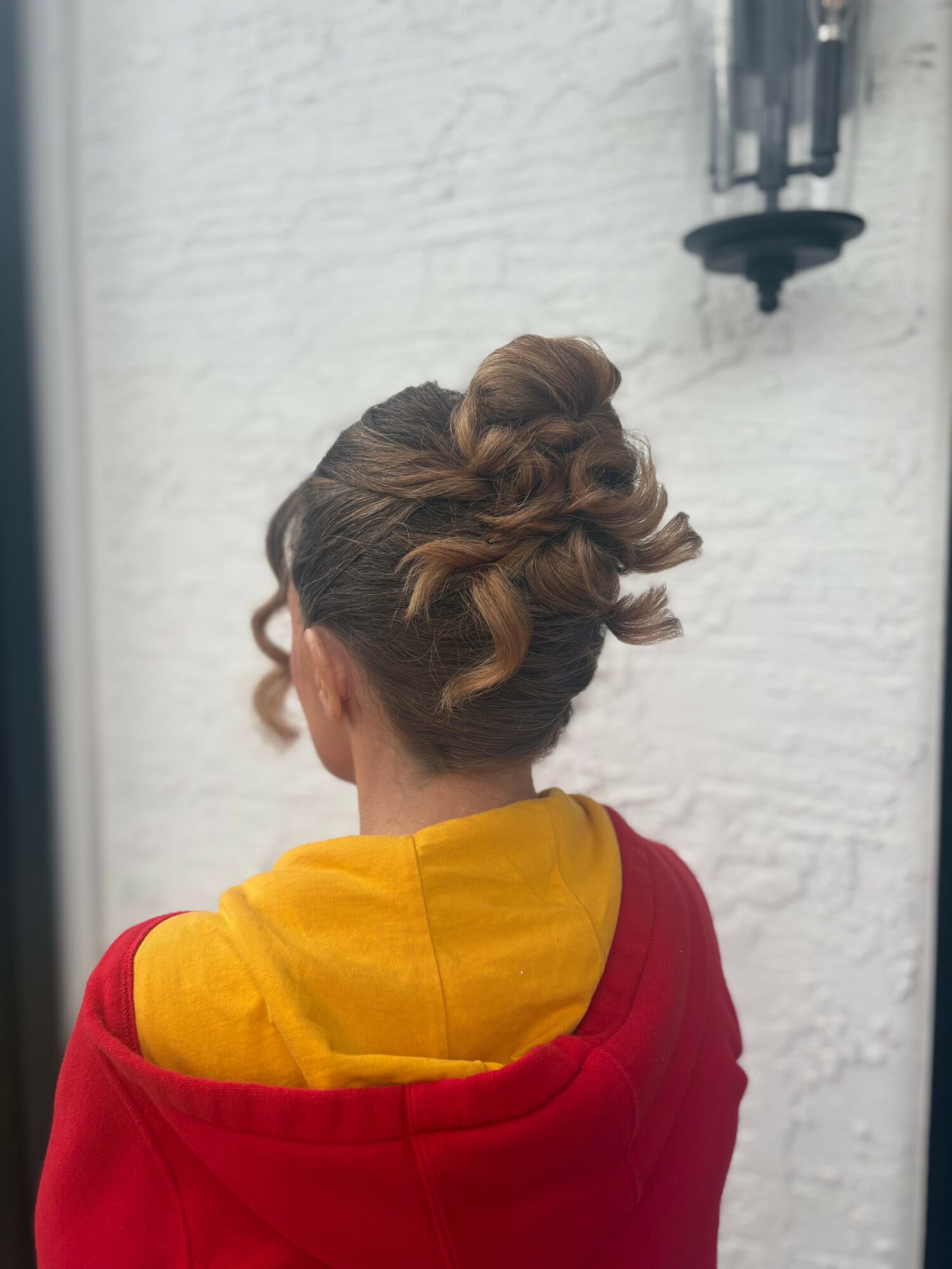 Back view of a woman with an intricate updo hairstyle, wearing a red and yellow garment, standing outdoors near a white wall.