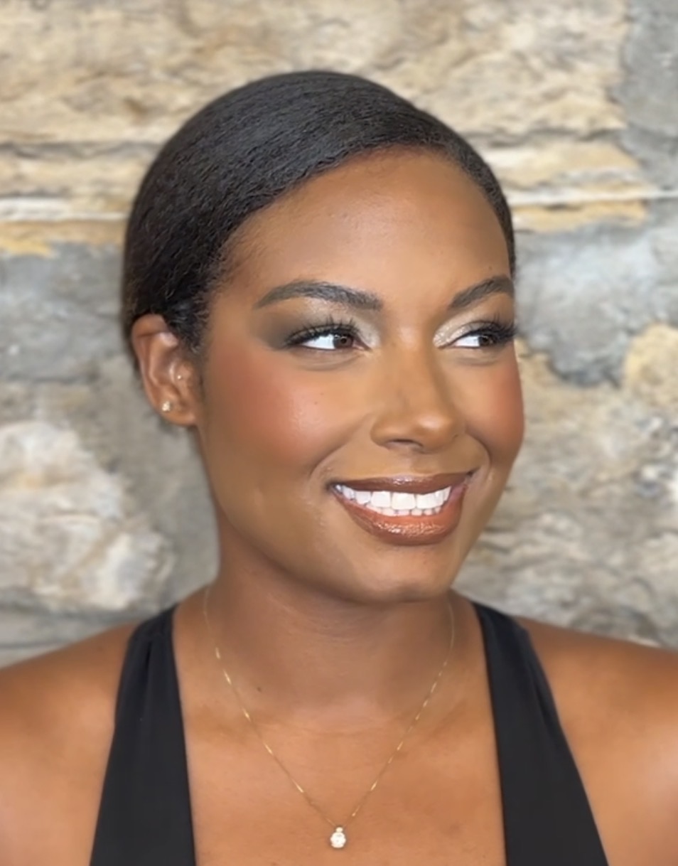 Smiling woman with short black hair, wearing a black top and a delicate necklace, against a stone wall background.