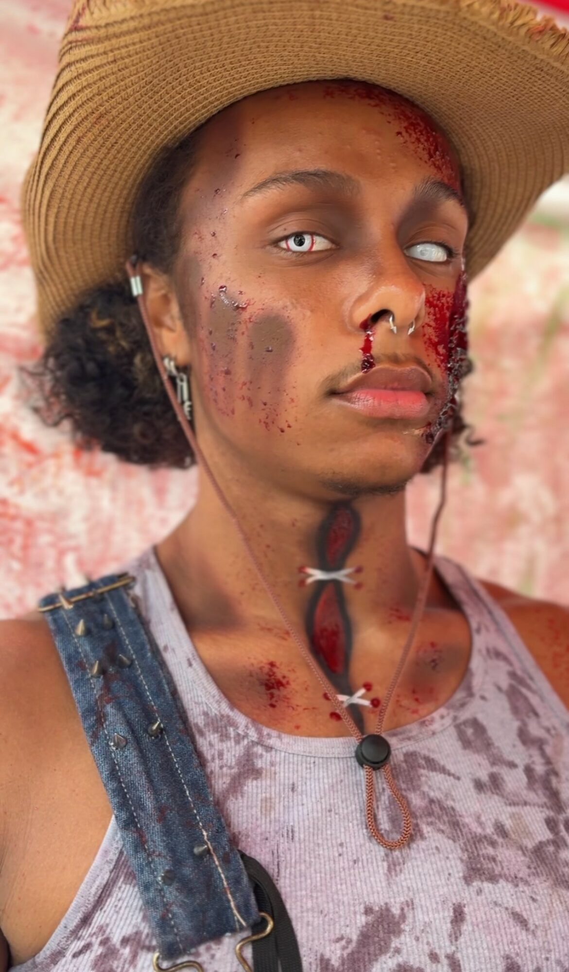 Person wearing a hat with blood and dirt on face and neck, wearing a tank top and denim strap, against a pink background.