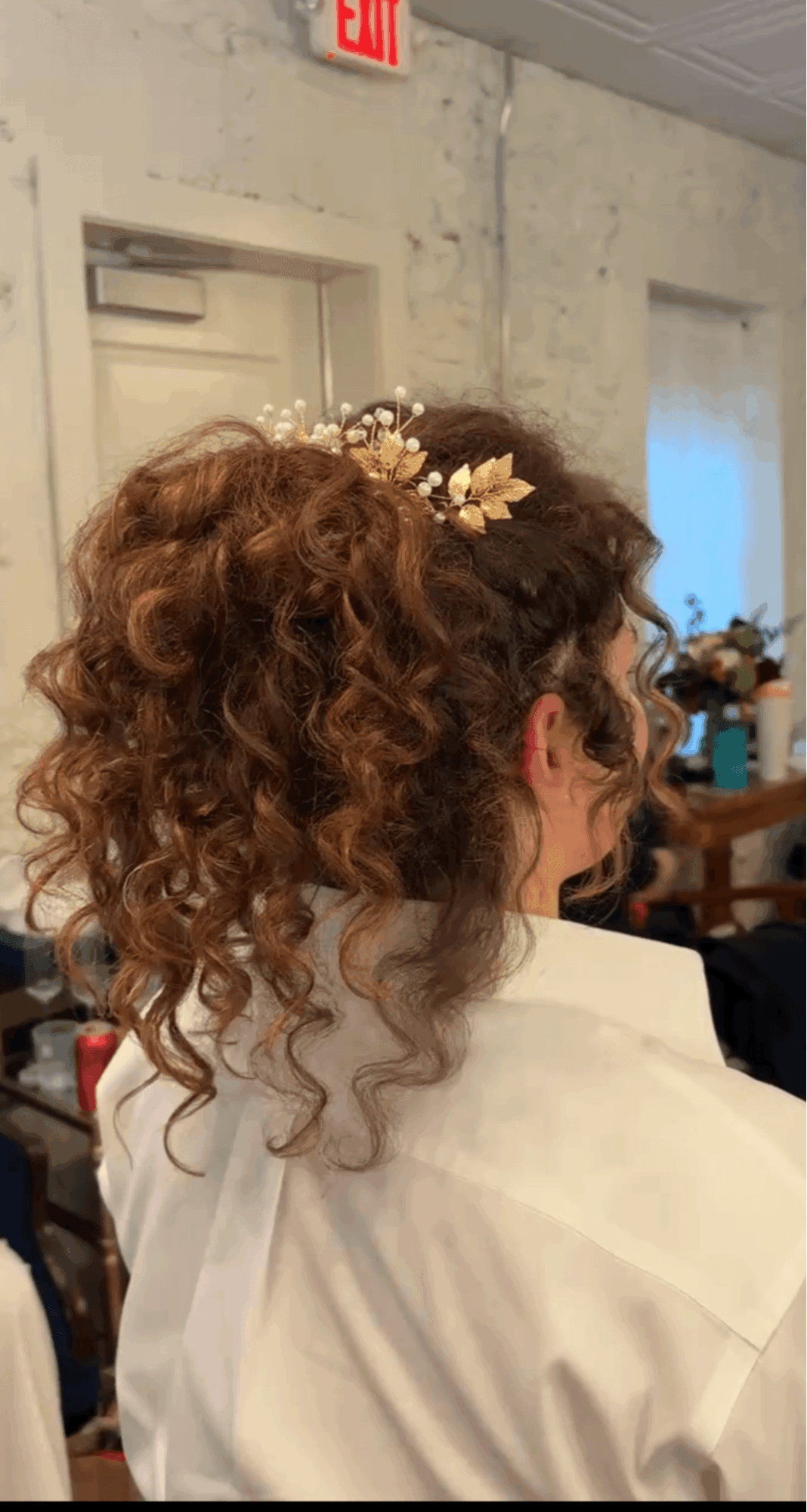 Person with curly hair styled up, wearing a white garment, in an indoor setting with a door and window in the background.
