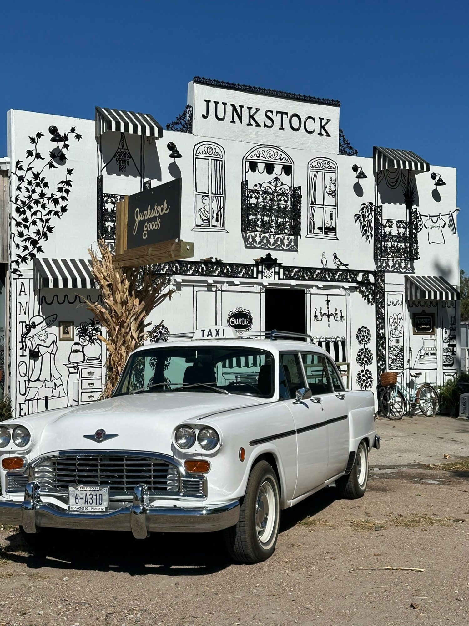 Vintage white car parked in front of a black and white building with signs reading 'Junkstock' and decorative elements.