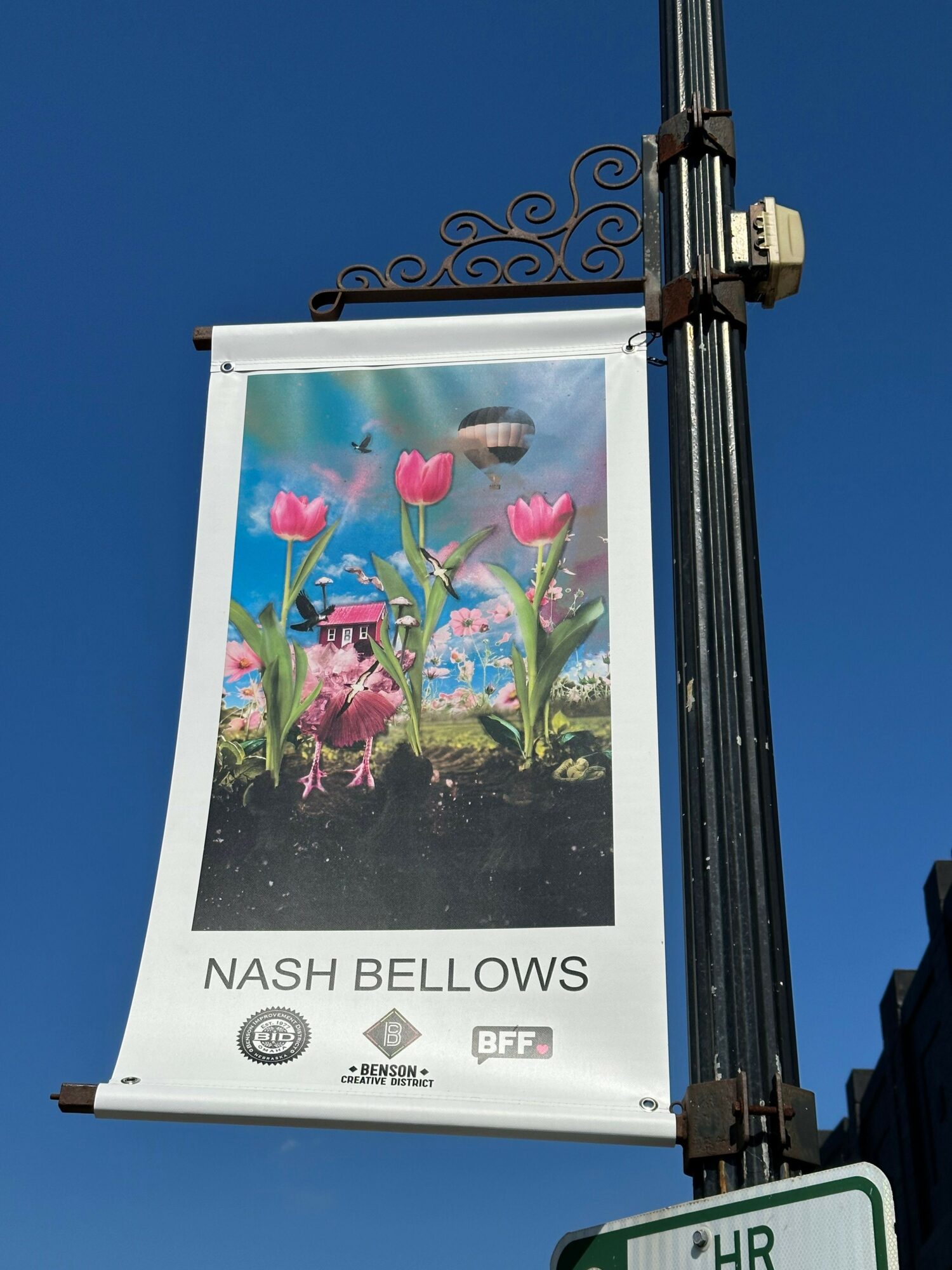Vertical banner with colorful floral design and text, mounted on a pole against a clear blue sky.