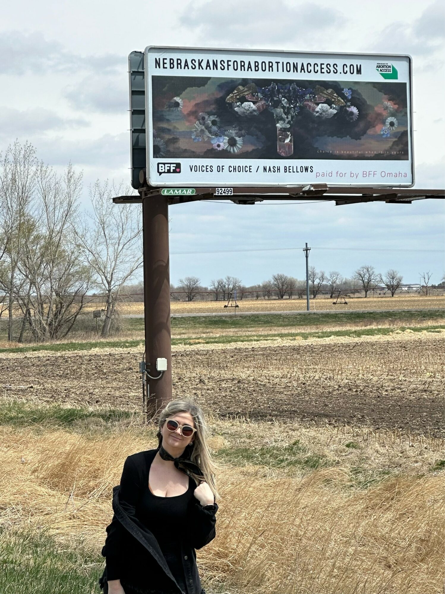 Woman with sunglasses standing outdoors near a billboard, landscape with fields and trees in background.