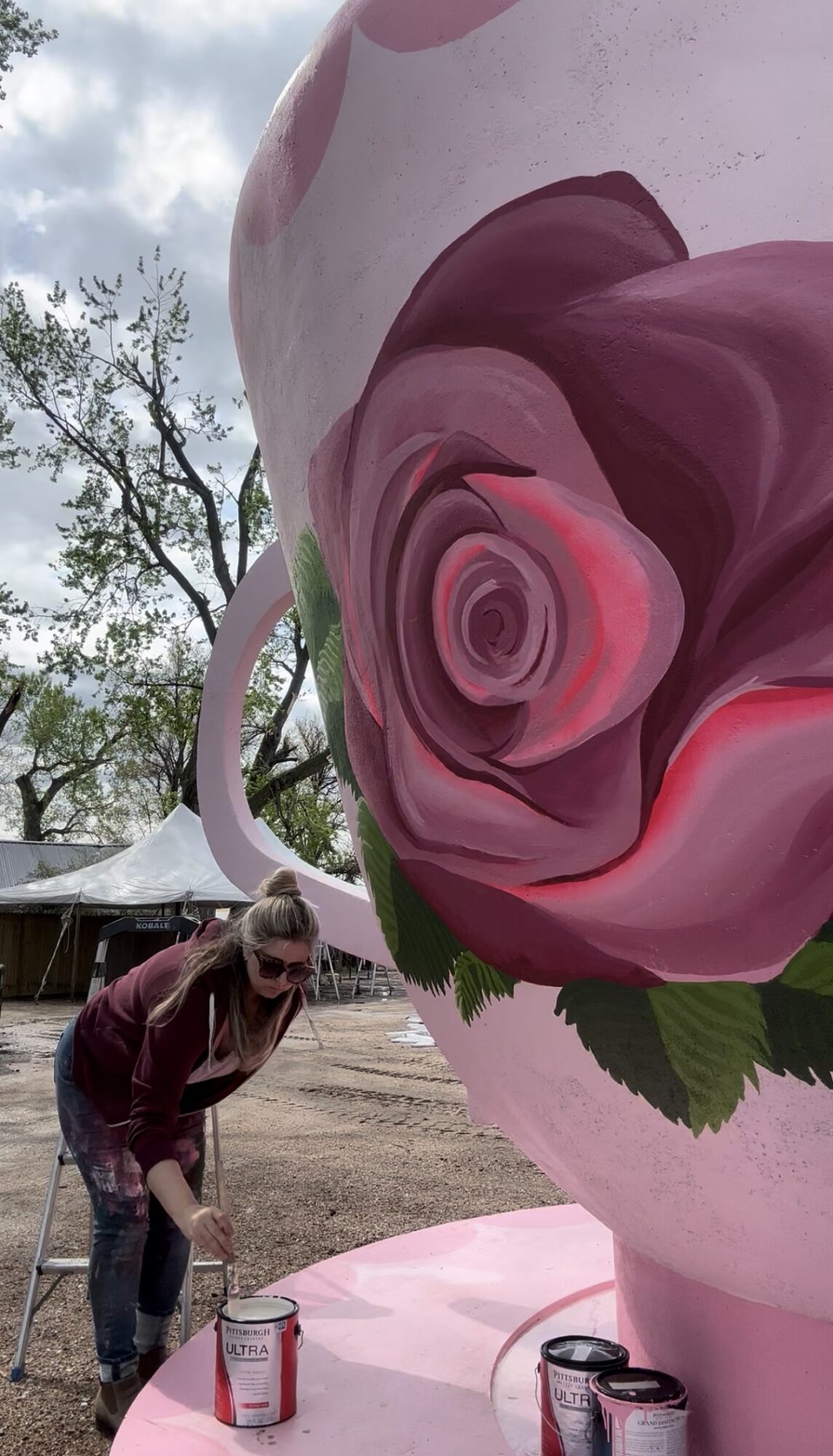 Person bending down near a large pink and purple rose sculpture outdoors, with trees and a tent in the background.