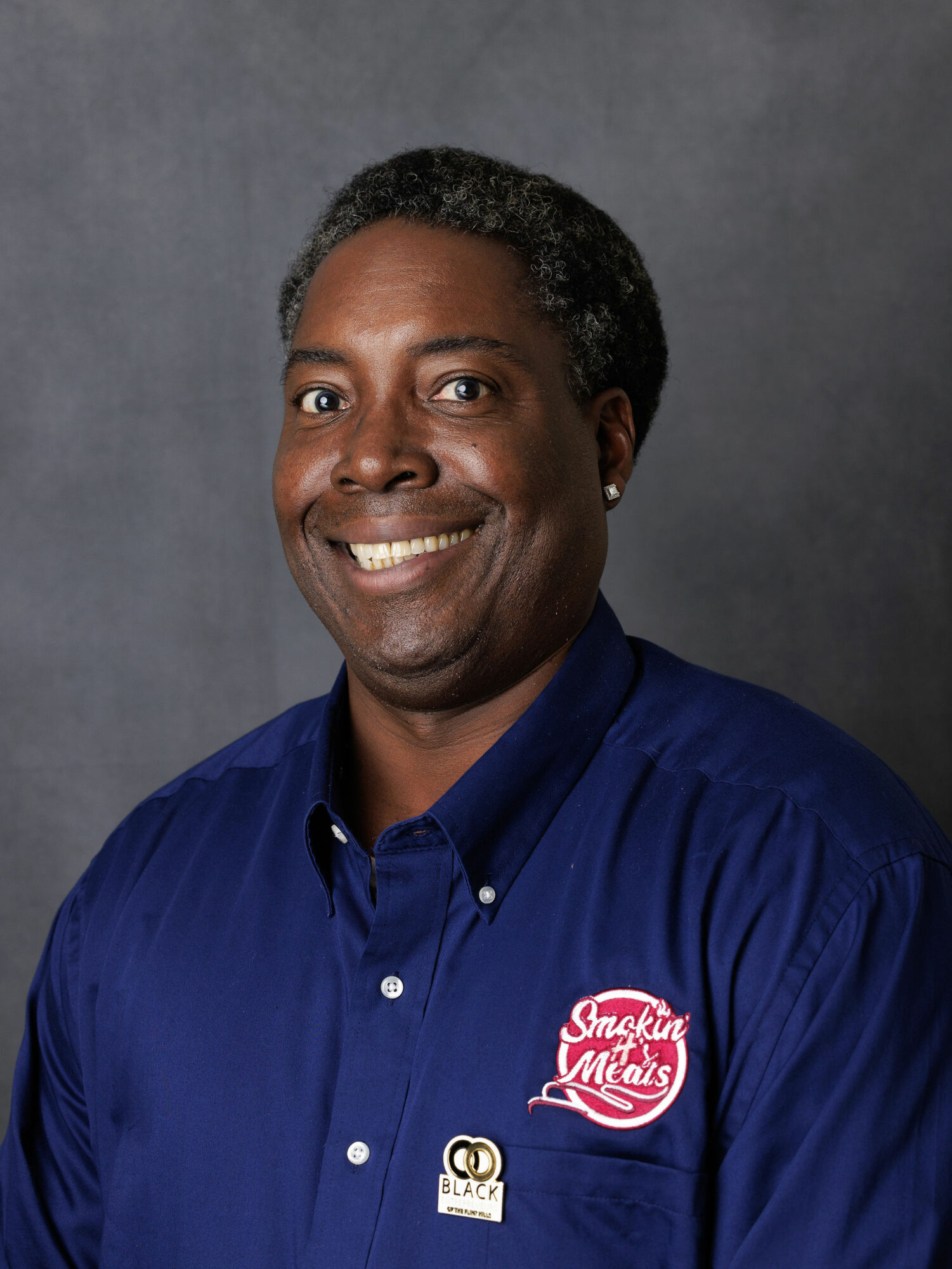 Smiling man with dark hair and dark skin wearing a blue shirt with a logo, against a gray background.