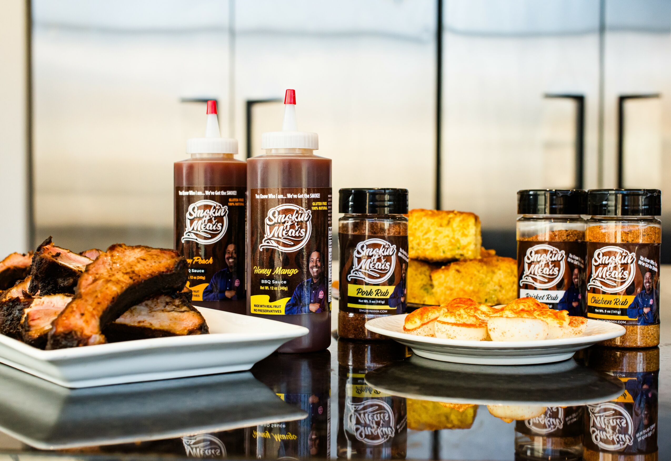 Assorted barbecue sauces, grilled chicken, cornbread, and sliced meat on plates, with a reflective background.