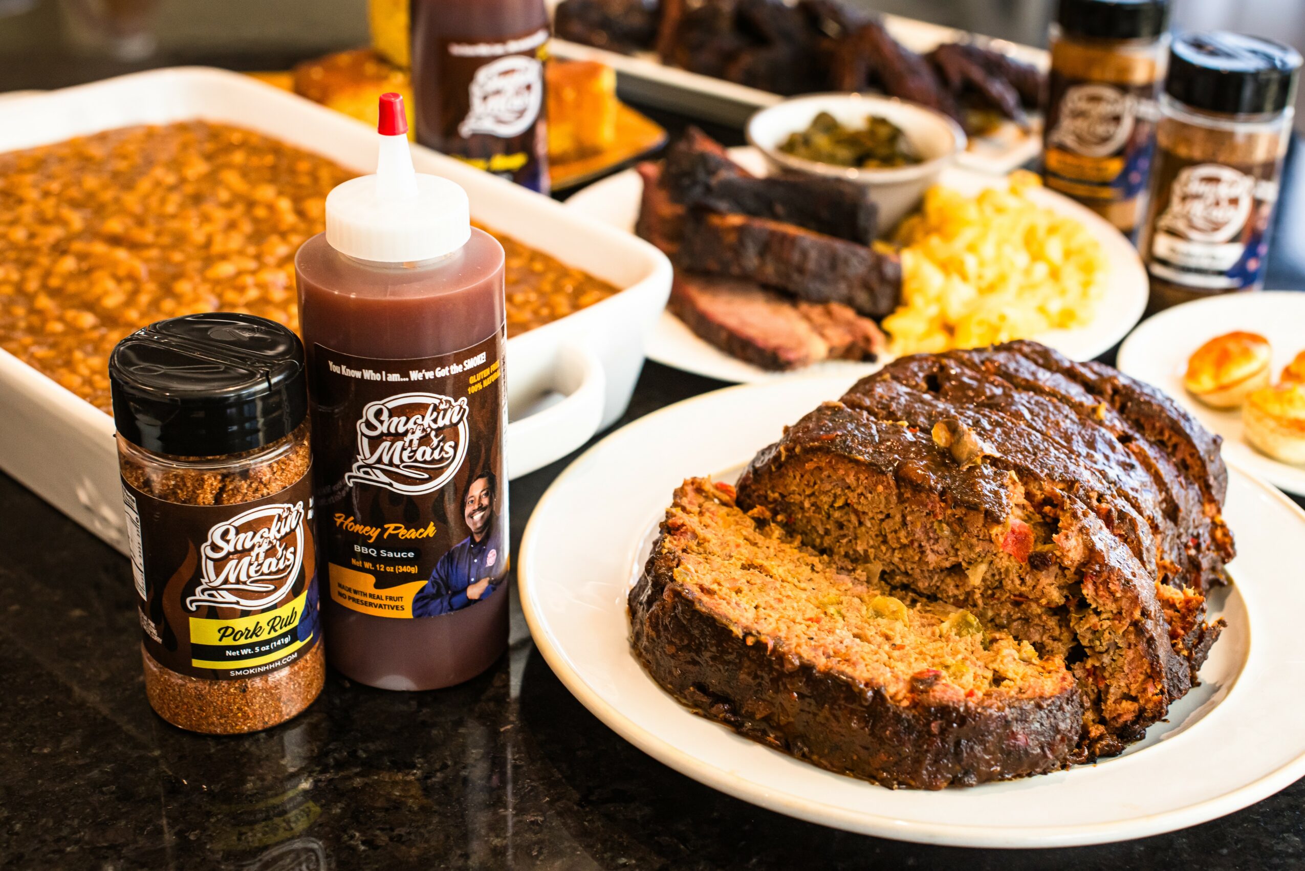 Plate with sliced meatloaf, surrounded by baked beans, mashed potatoes, and condiments, on a black countertop.