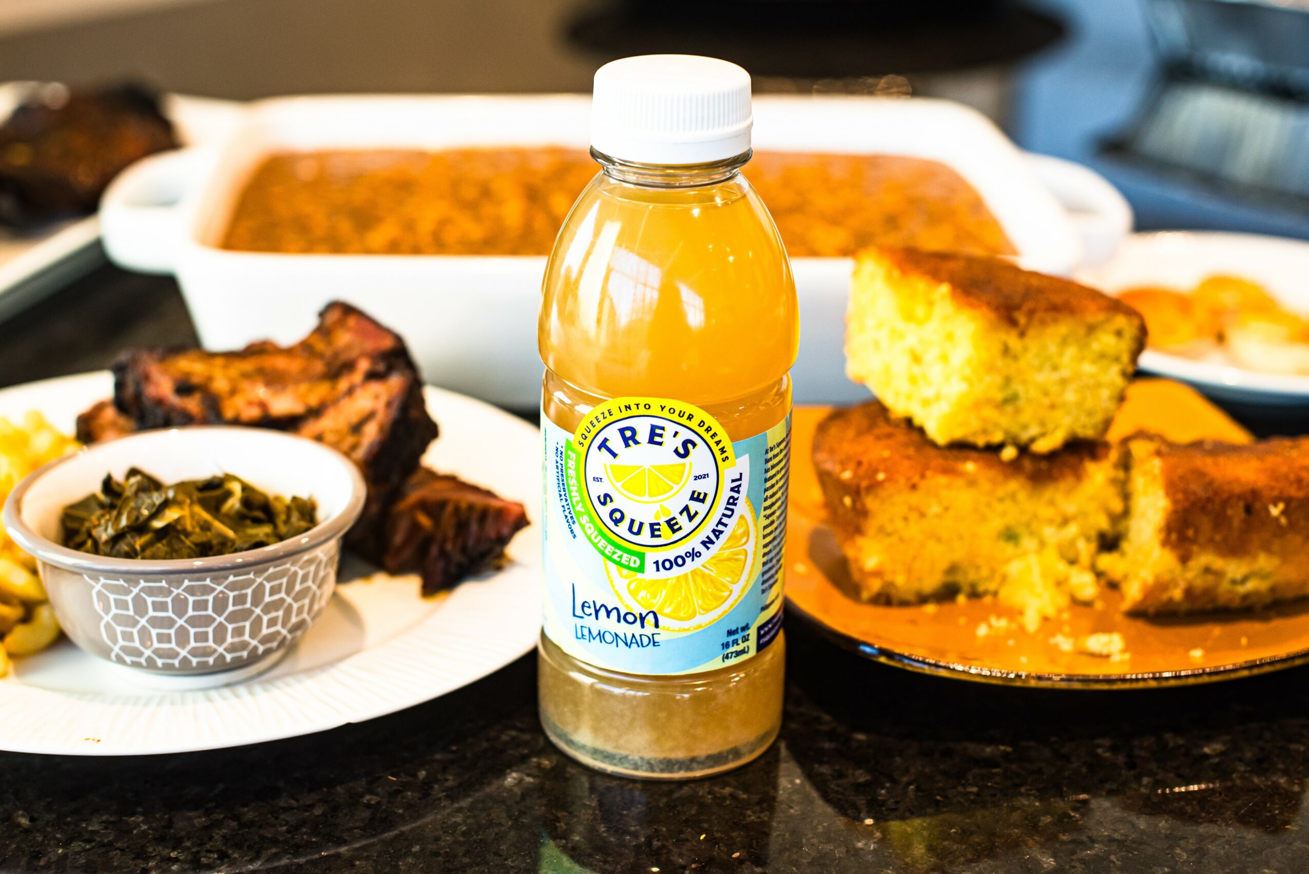 Bottle of lemon-flavored beverage in front of baked goods and cooked dishes on a table.