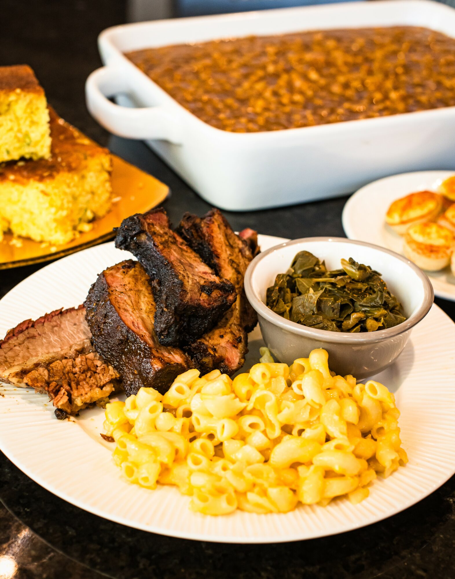 Plate with grilled meat, macaroni, and greens, with baked cornbread and a casserole dish in the background.