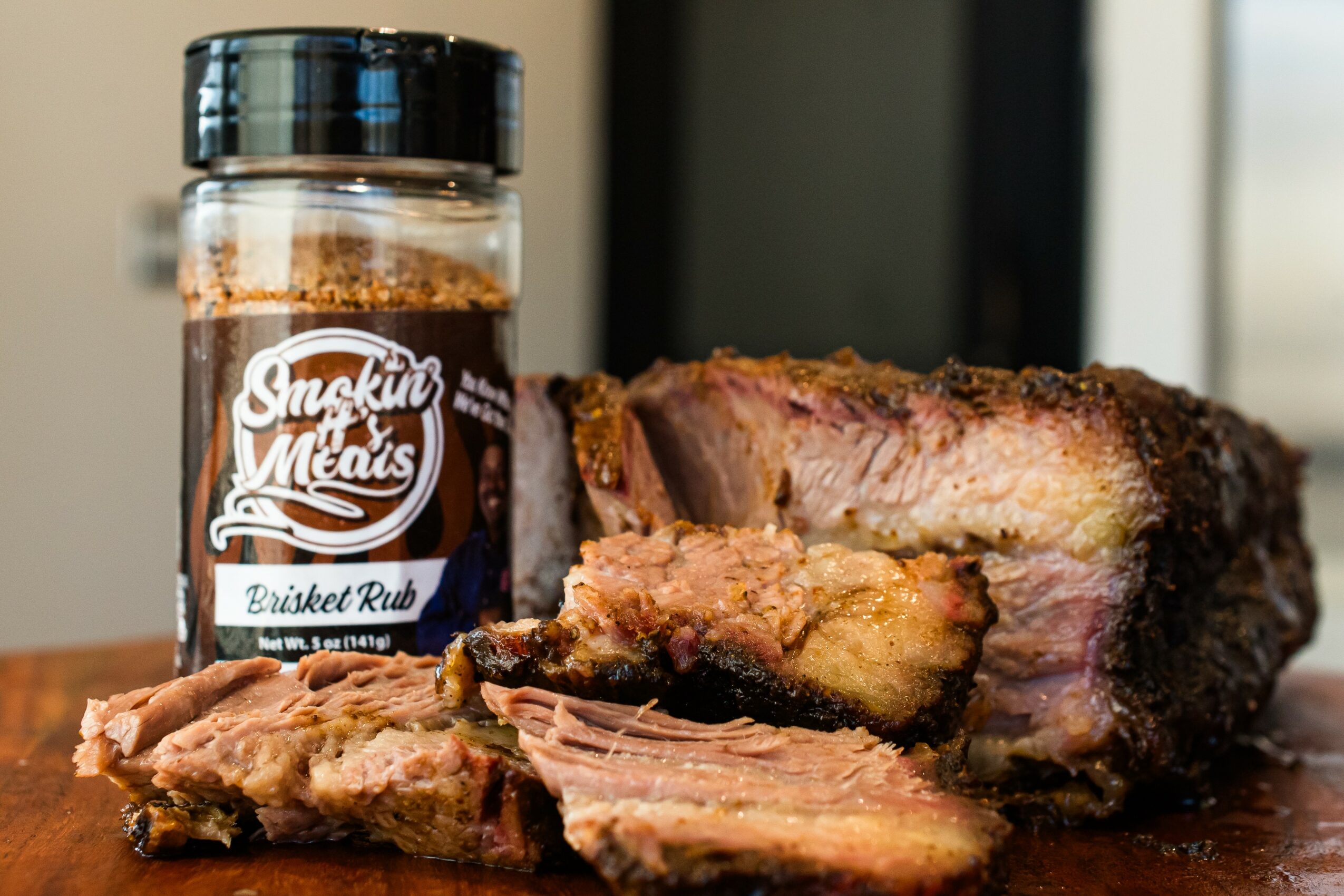 Cooked brisket with slices on a cutting board, jar of brisket rub in background, indoors, natural light, close-up