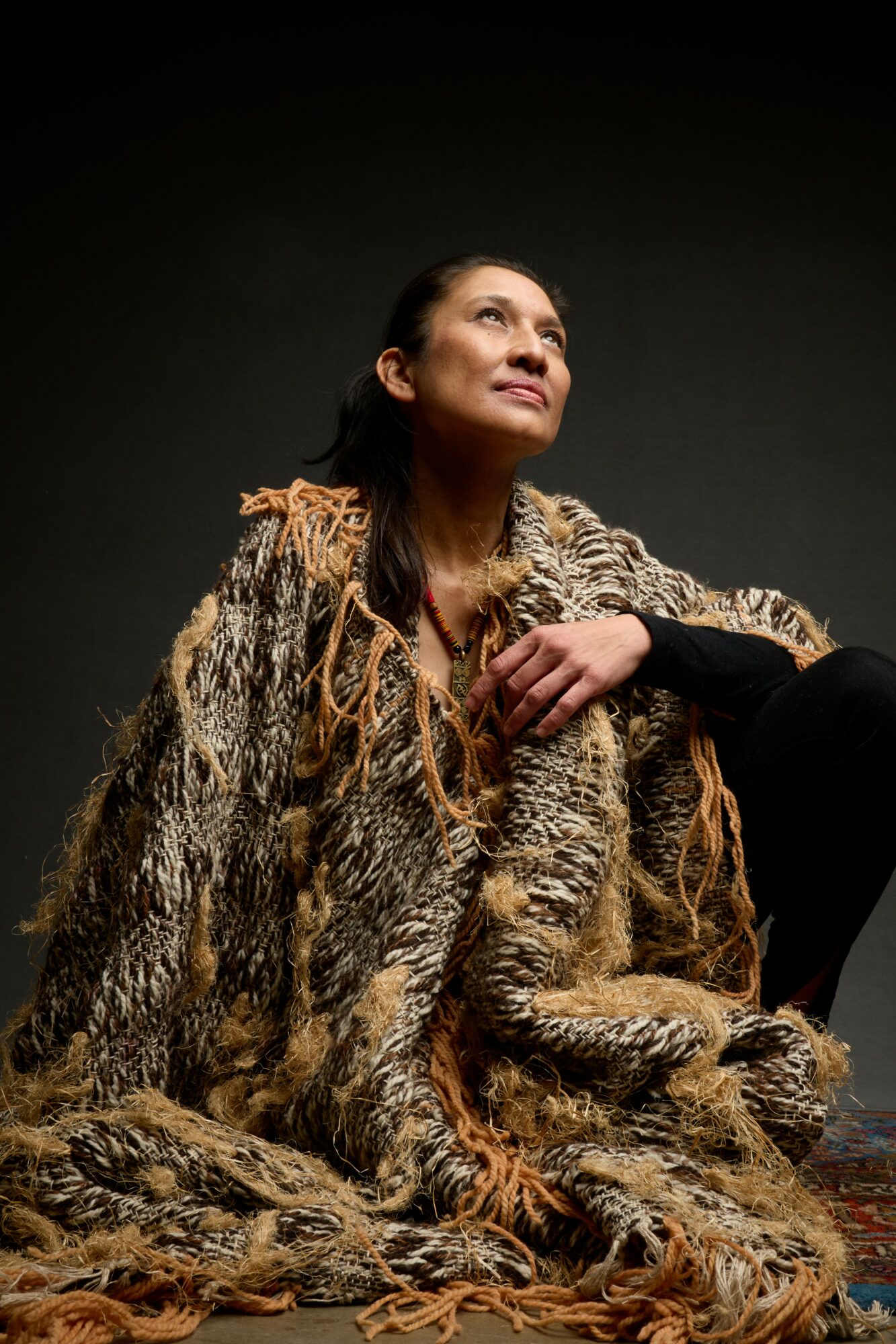 Woman wearing a fur coat, looking upward with a neutral expression, against a dark background.
