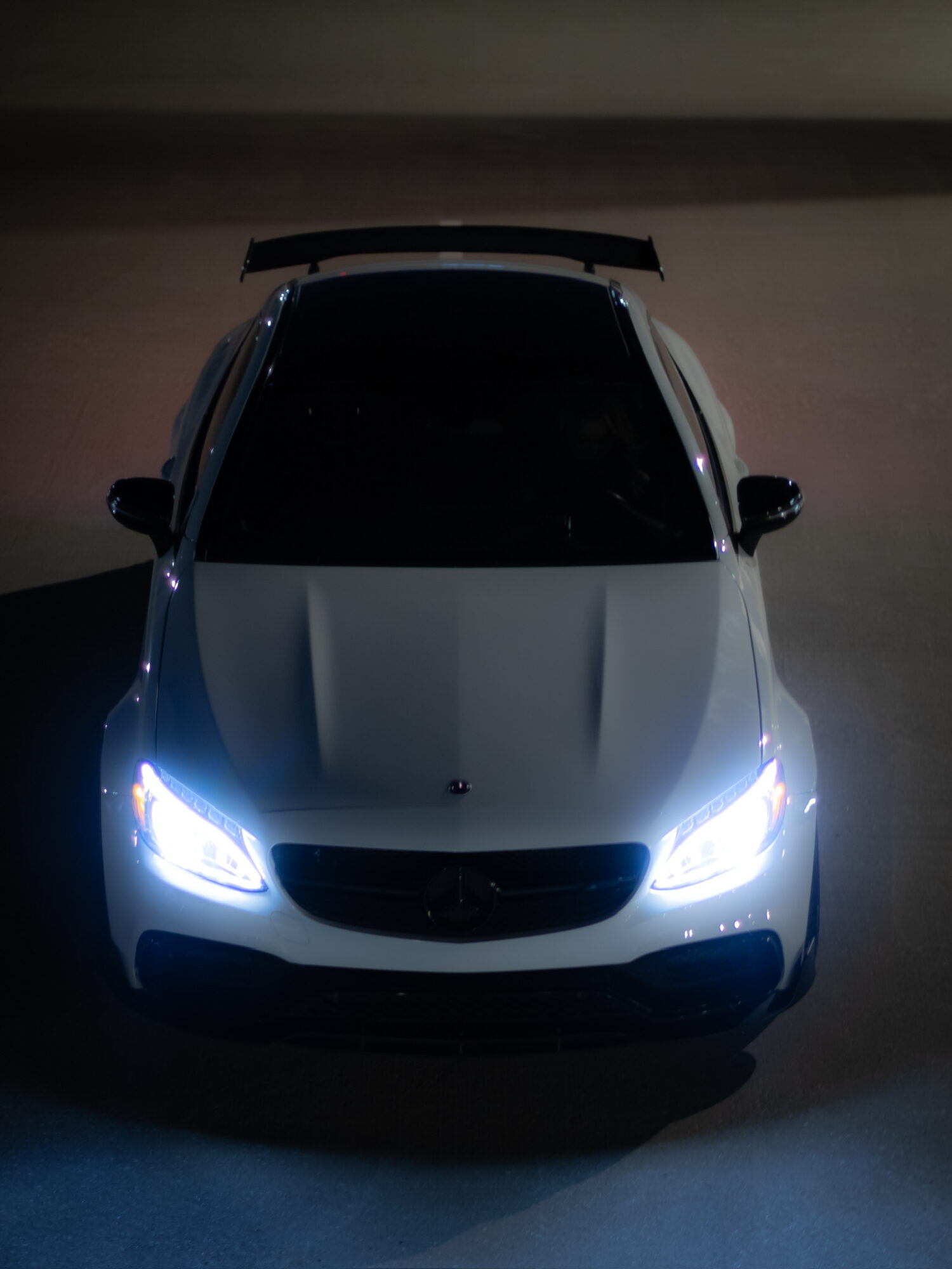 Front view of a white car with headlights on, parked indoors, with a spoiler on the rear.