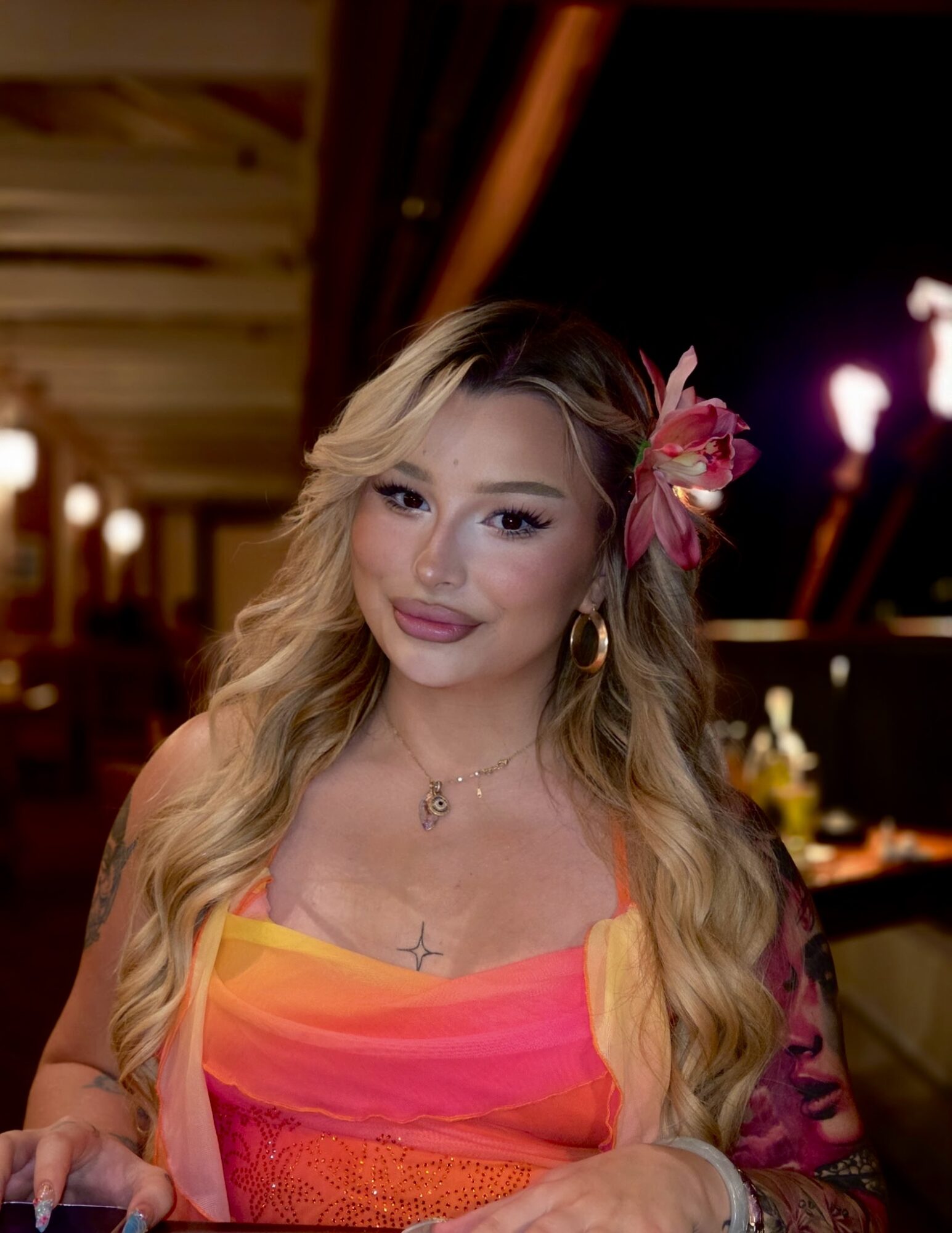 Woman with long wavy blonde hair, wearing a pink and orange dress, and a pink flower in her hair, in a dimly lit setting.