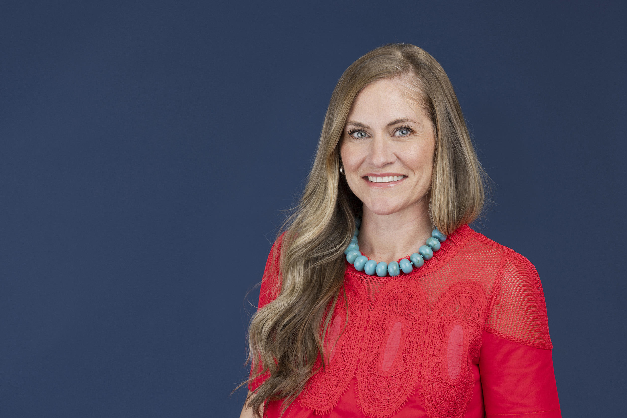 Woman with long wavy hair wearing a red top and turquoise necklace, smiling, against a dark blue background.