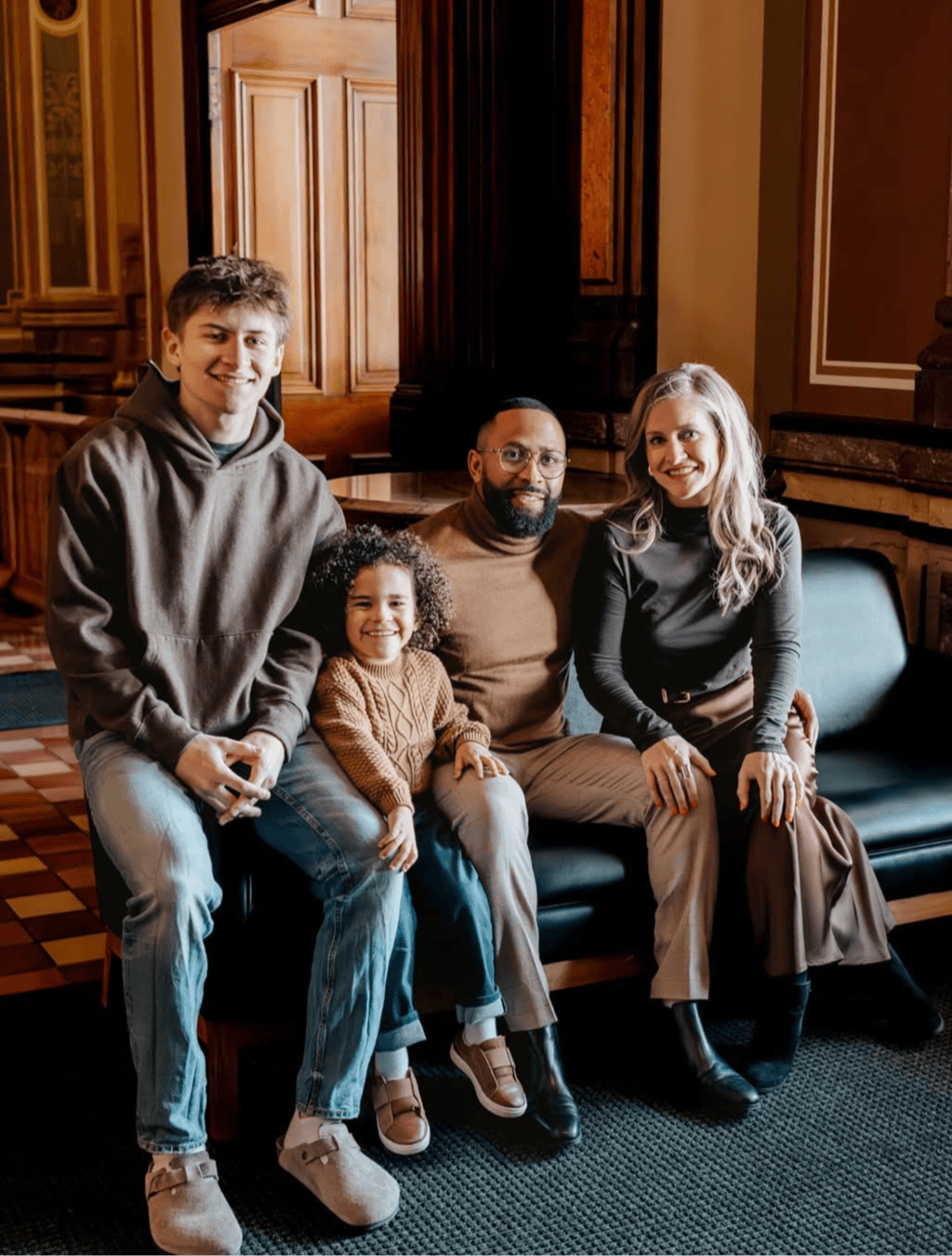 Four people sitting on a sofa in a room with wooden paneling and large windows, smiling at the camera.