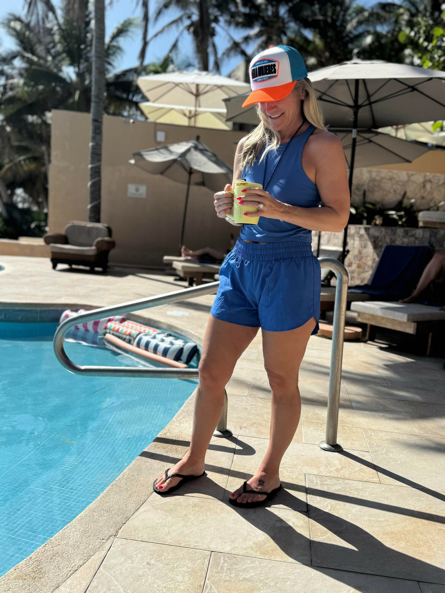 Woman in blue shorts and tank top holding a drink by a pool with umbrellas and lounge chairs, sunny outdoor setting.